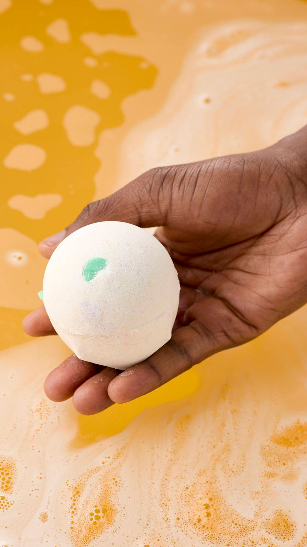A model's hand holds a spherical, off-white "Dragon's Egg" bath bomb with small slivers of colour over a tub of swirling orange bathwater. The water is frothy with many small bubbles on the surface.