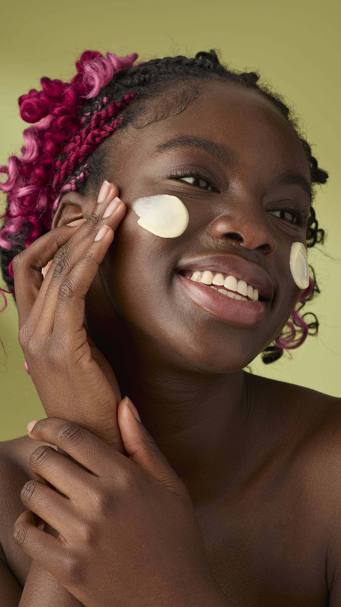 A woman with pink and black braids smiles as she applies a smooth, ivory Oat facial cleanser to her cheeks. The thick, creamy product stands out against her skin and the solid olive-green background, emphasising its rich texture.