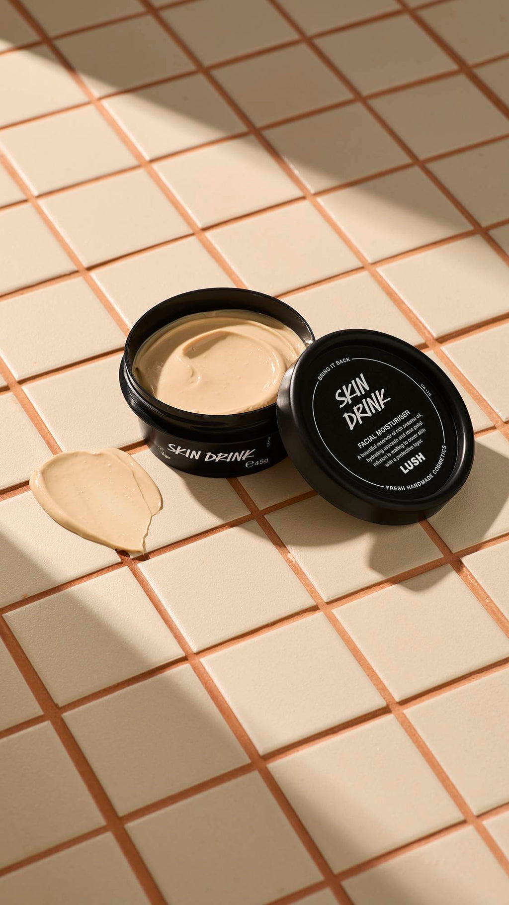 A black pot of Lush Skin Drink facial moisturizer resting on a beige tiled surface with the lid leaning against the container, next to a smear of the cream.