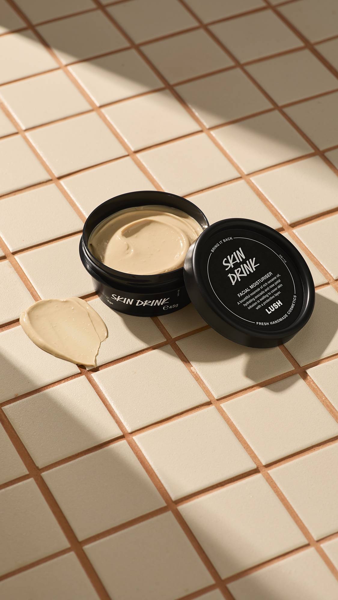 A black pot of Lush Skin Drink facial moisturizer resting on a beige tiled surface with the lid leaning against the container, next to a smear of the cream.