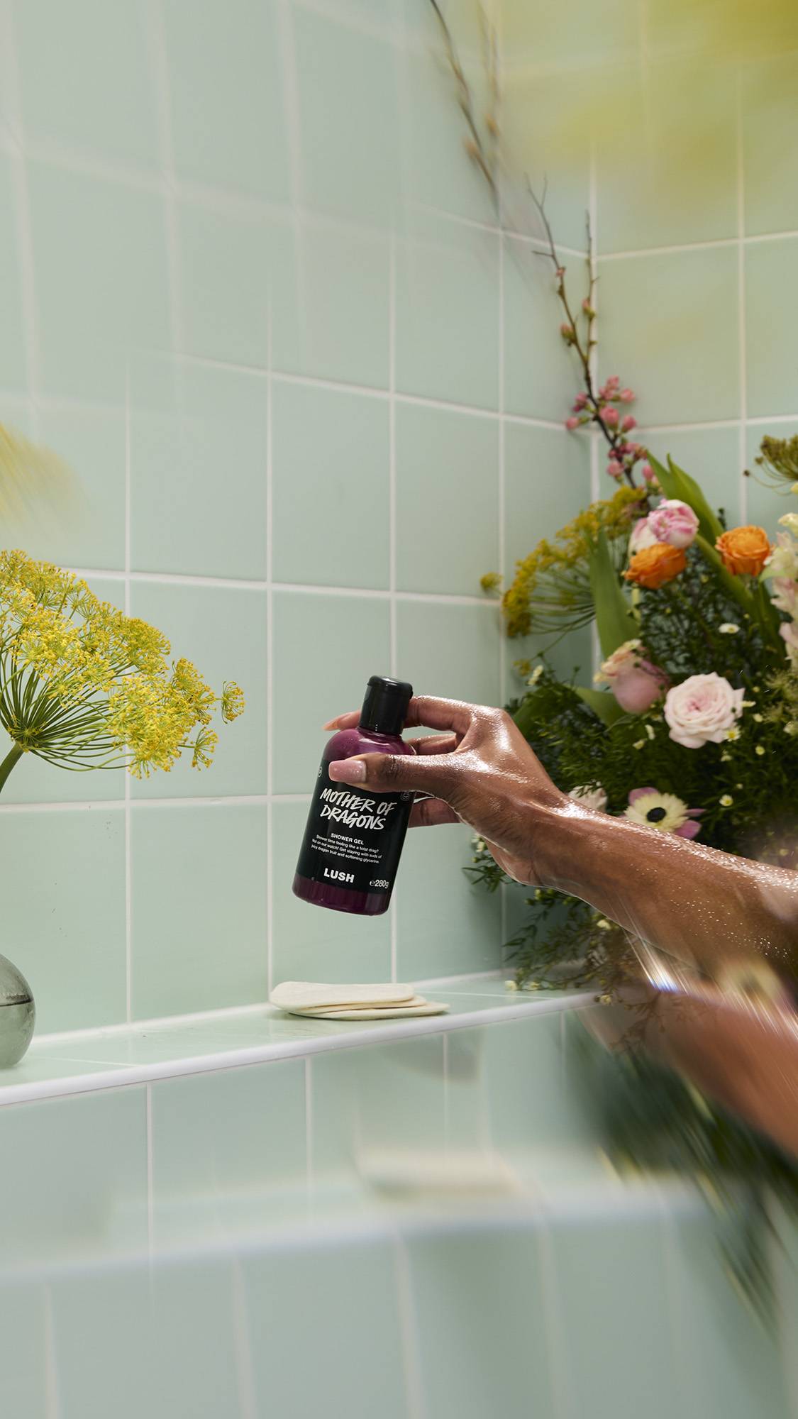 A bottle of purple, shimmery "Mother of Dragons" Shower Gel sits on a white tiled shelf. The label mentions "juicy dragon fruit and softening glycerine." Bubbles and greenery surround the bottle against a mint green tiled background.