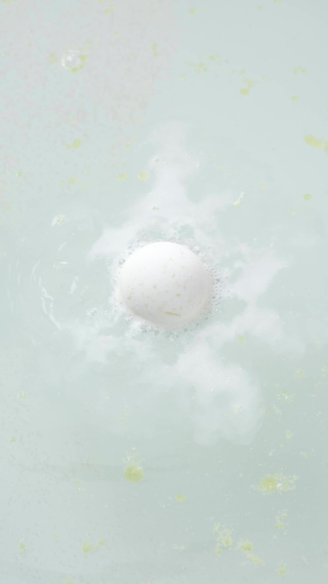 The white, spherical Butterball bath bomb sits on a plain, light gray background. The product features a matte, slightly textured surface with small, visible brown flecks of cocoa butter throughout.