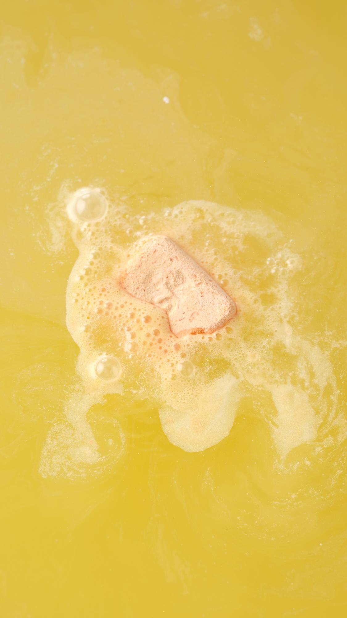 An aerial view of the Cold Water Soother bath bomb dissolving. The product creates a thick layer of white velvety foam that ripples and swirls across the surface of yellow water.