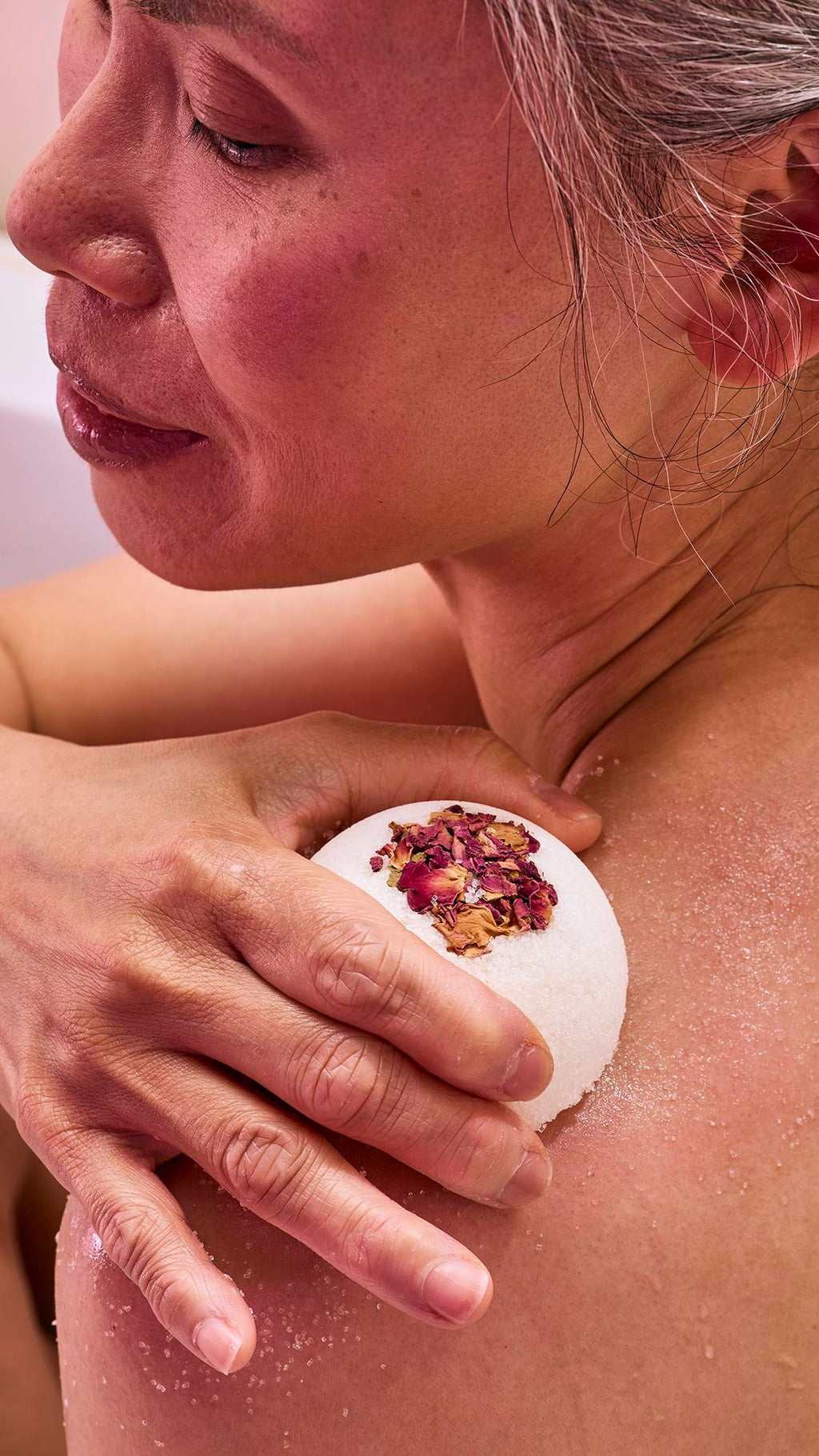A model in a bath uses a white, dome-shaped "Comforter Rose" solid body scrub. They are rubbing the textured surface against their shoulder, leaving a fine, white exfoliating residue on their skin.