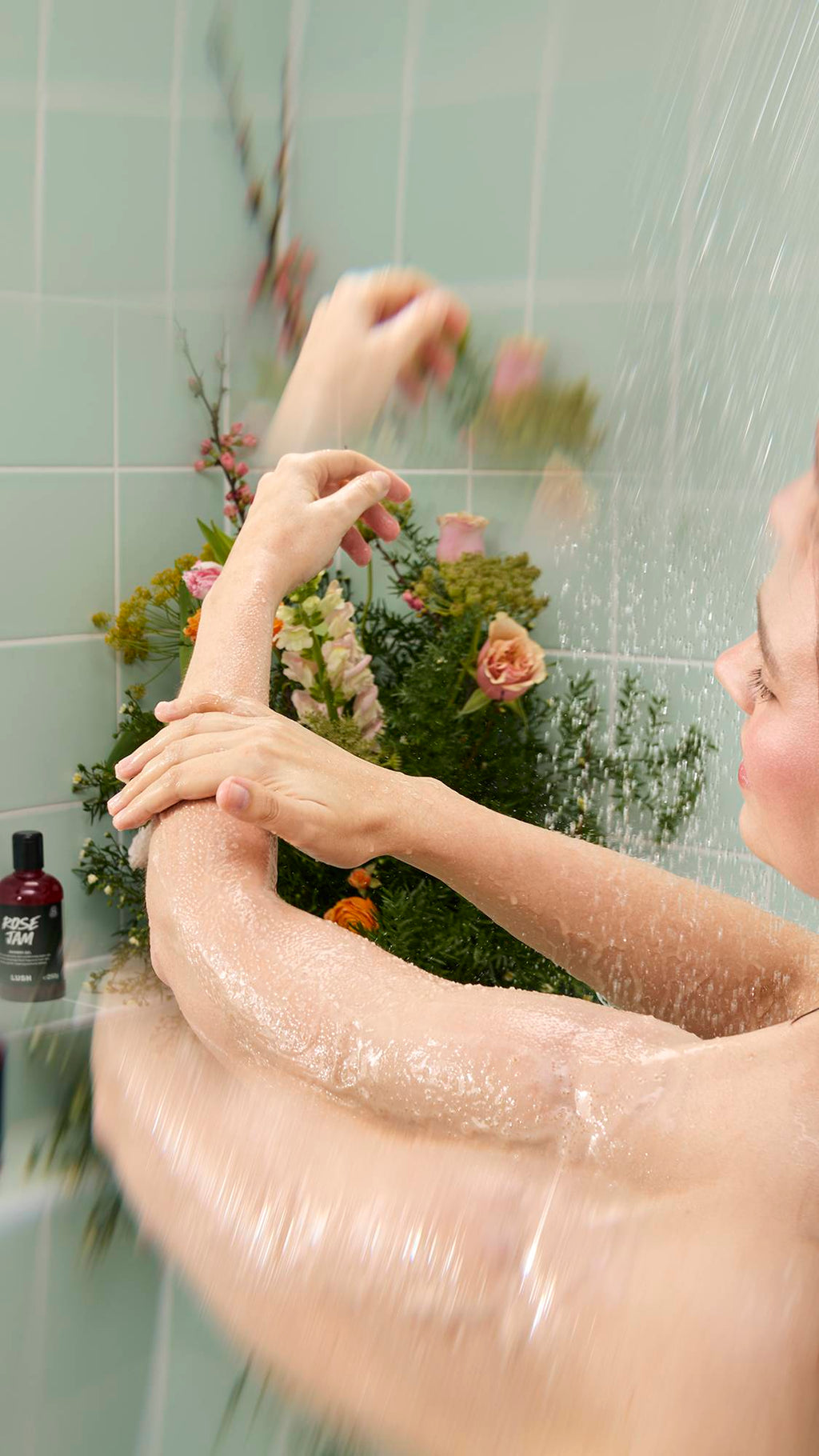 A person stands under a shower stream against a background of mint green tiles and flowers, massaging Crème Brûlée Body Scrub into their wet arm. A bottle of "Rose Jam" Shower Gel is visible on the shelf in the background.