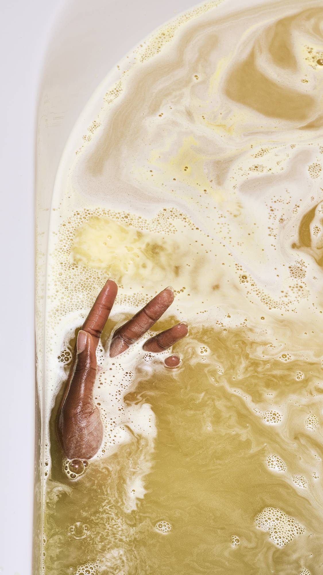 A close-up of a model's hands submerged in a golden bath. Thick, creamy yellow foam swirls across the water's surface as a bath bomb dissolves, creating a marbled pattern of light and dark gold.