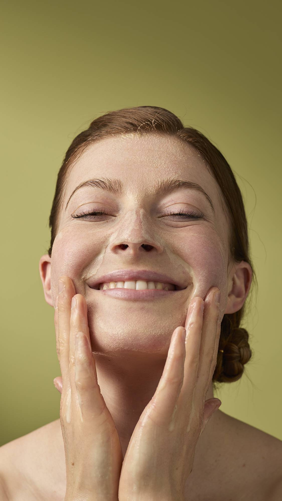 A person with their eyes closed and a gentle smile massages a foamy cleanser into their face. Their wet hands are covered in light soap suds, and they stand against a solid olive-green background.