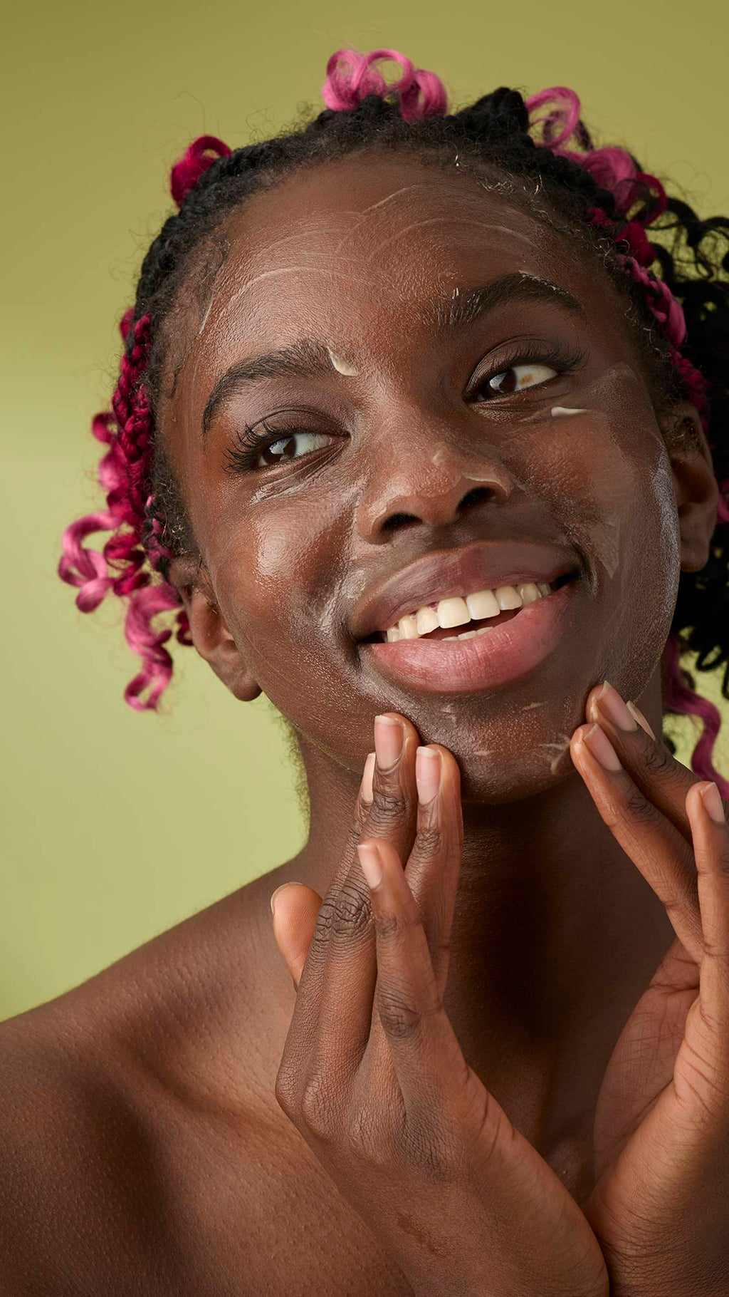 A person with pink braids smiles while gently massaging Ultrabland cleanser onto their jawline and cheeks against an olive green background.