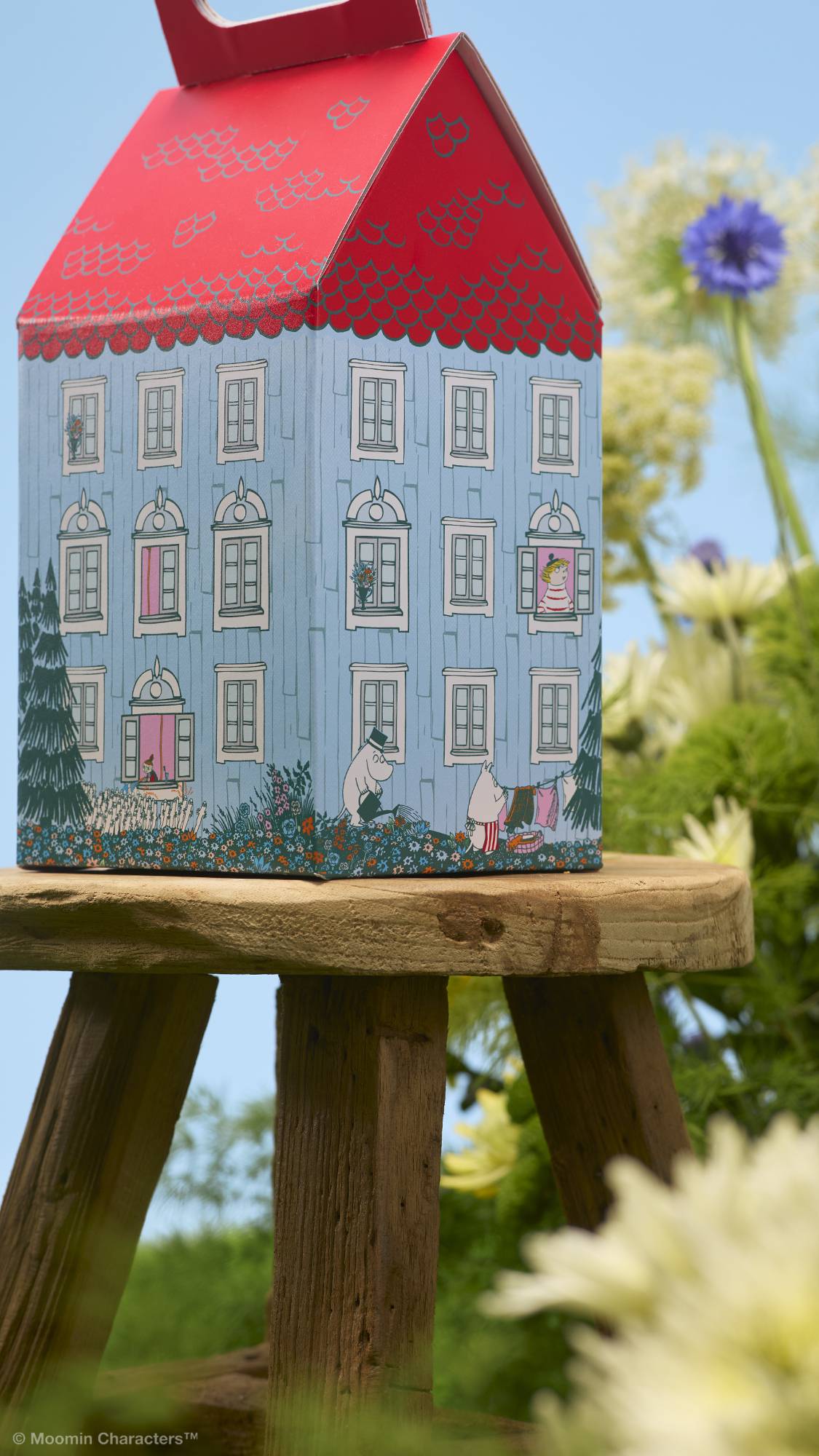 A close-up shot of a house-shaped gift box resting on a rustic wooden stool. The blue box features detailed illustrations of Moomin characters in the garden and windows. Out-of-focus green foliage and wildflowers surround it under a blue sky.