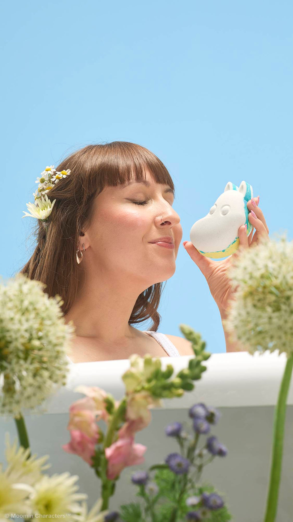 A close-up shot of a person with flowers in their hair, smiling with closed eyes as they bring a white Moomintroll bath bomb up to their nose to smell it. Wildflowers are in the blurry foreground against a clear blue sky.