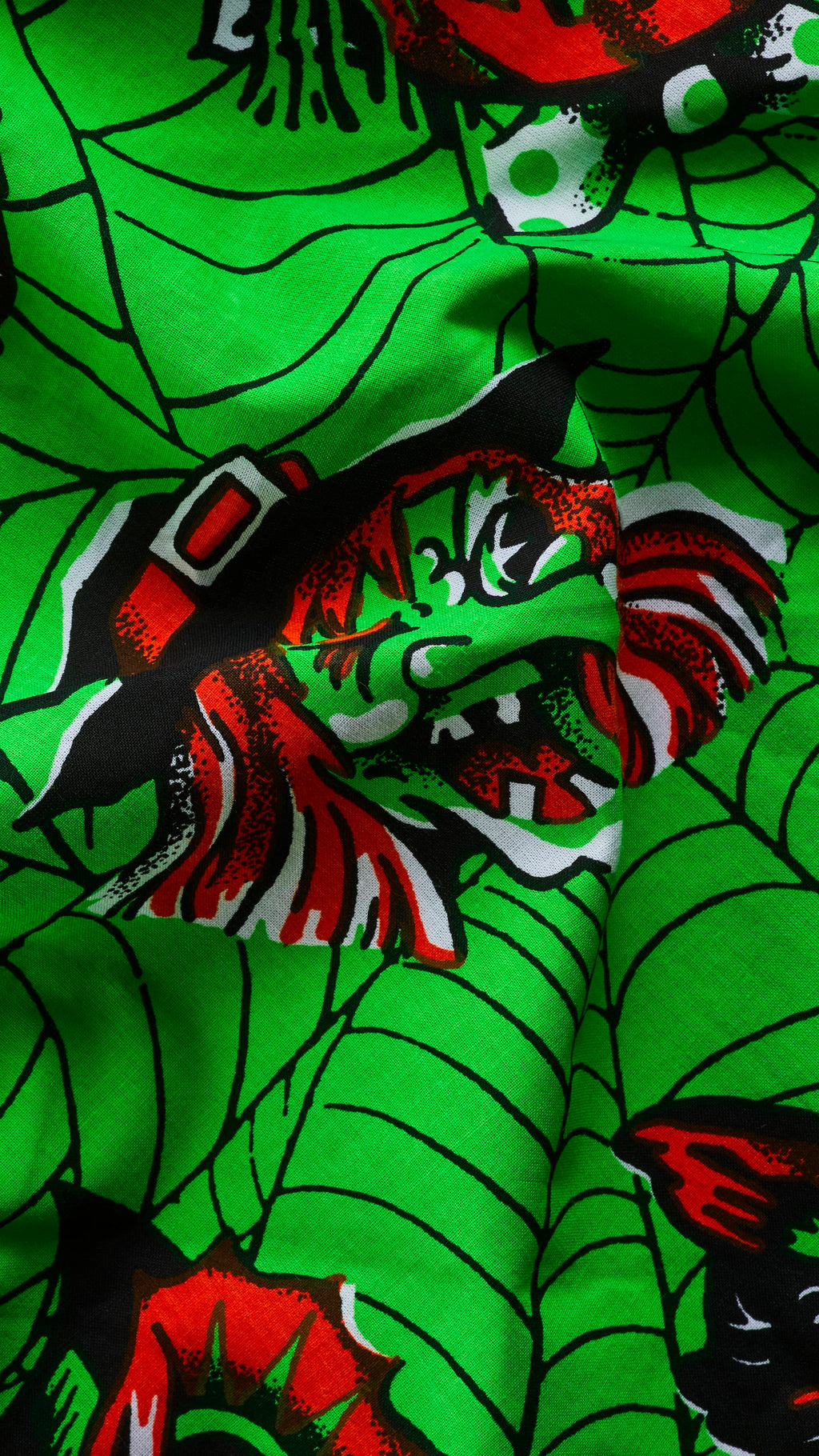 A close-up of a rumpled bright green fabric knot wrap with a black cobweb pattern. The fabric features a vintage-style illustration of a green-faced witch wearing a black and red pointy hat with a buckle.