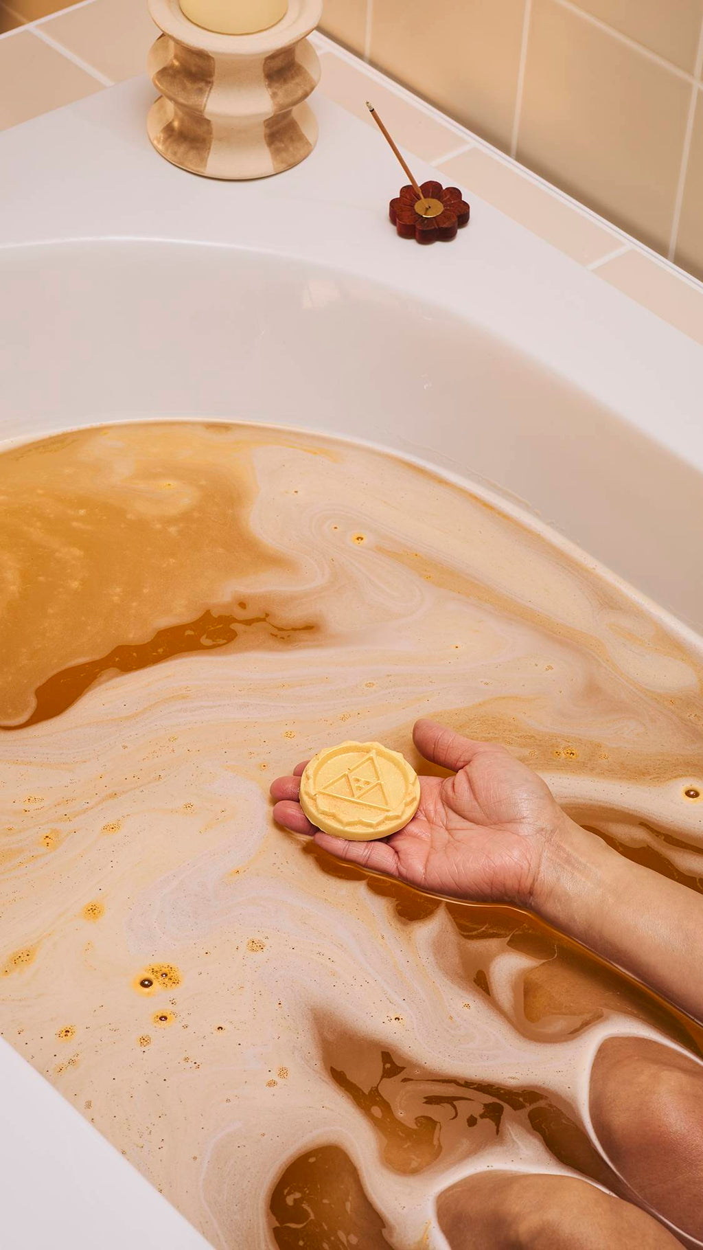 A person rests in a bath filled with amber-coloured, foamy water. They hold a circular yellow solid body oil embossed with a triangle design in their palm. A lit candle and incense stick sit on the bath's edge against neutral-toned tiles.