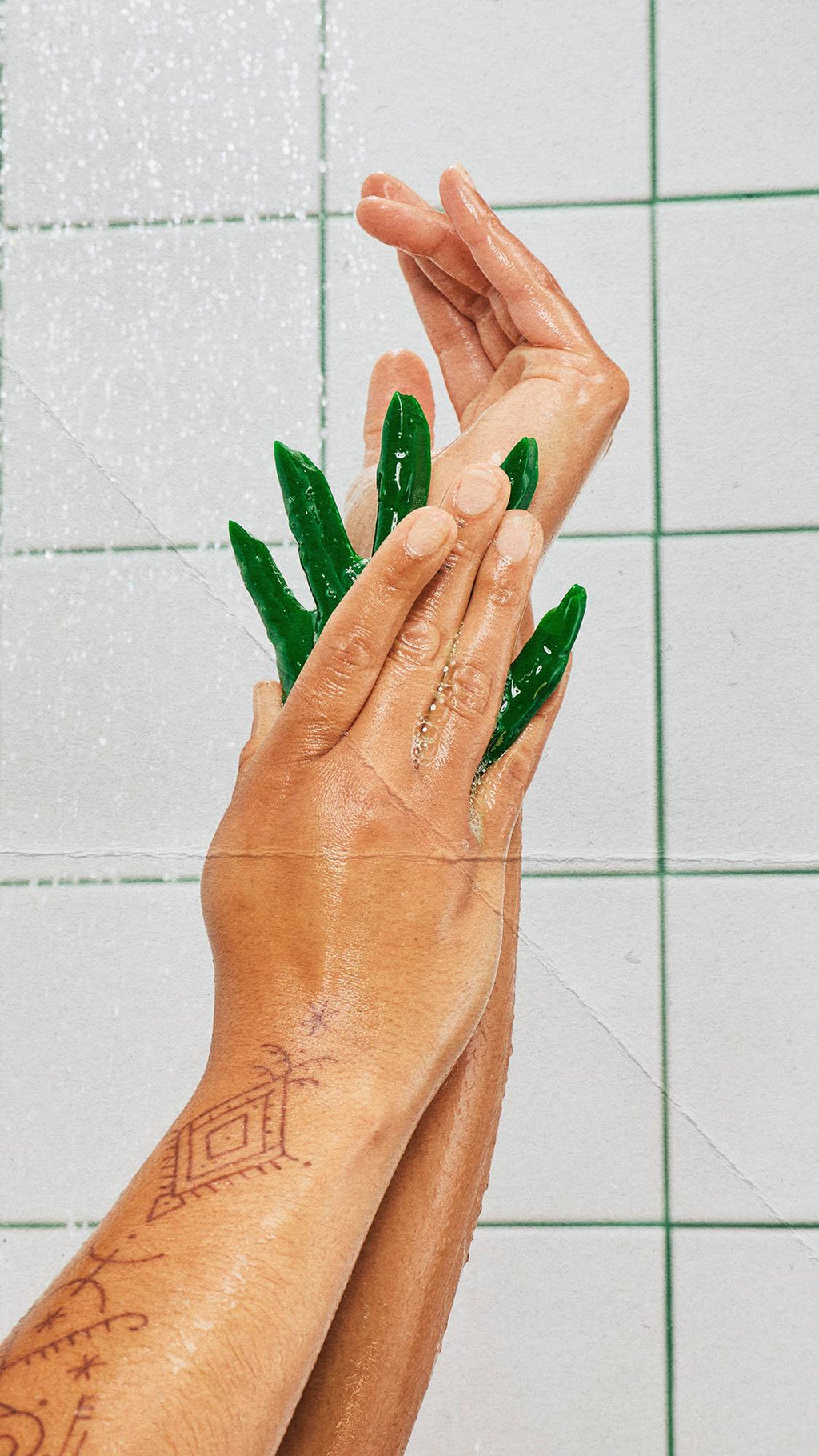 Two tattooed hands lathering a dark green, witch's hand-shaped shower gummy under streaming shower water. The wet hands are crossed, rubbing the product together. White square wall tiles with green grout lines form the background.