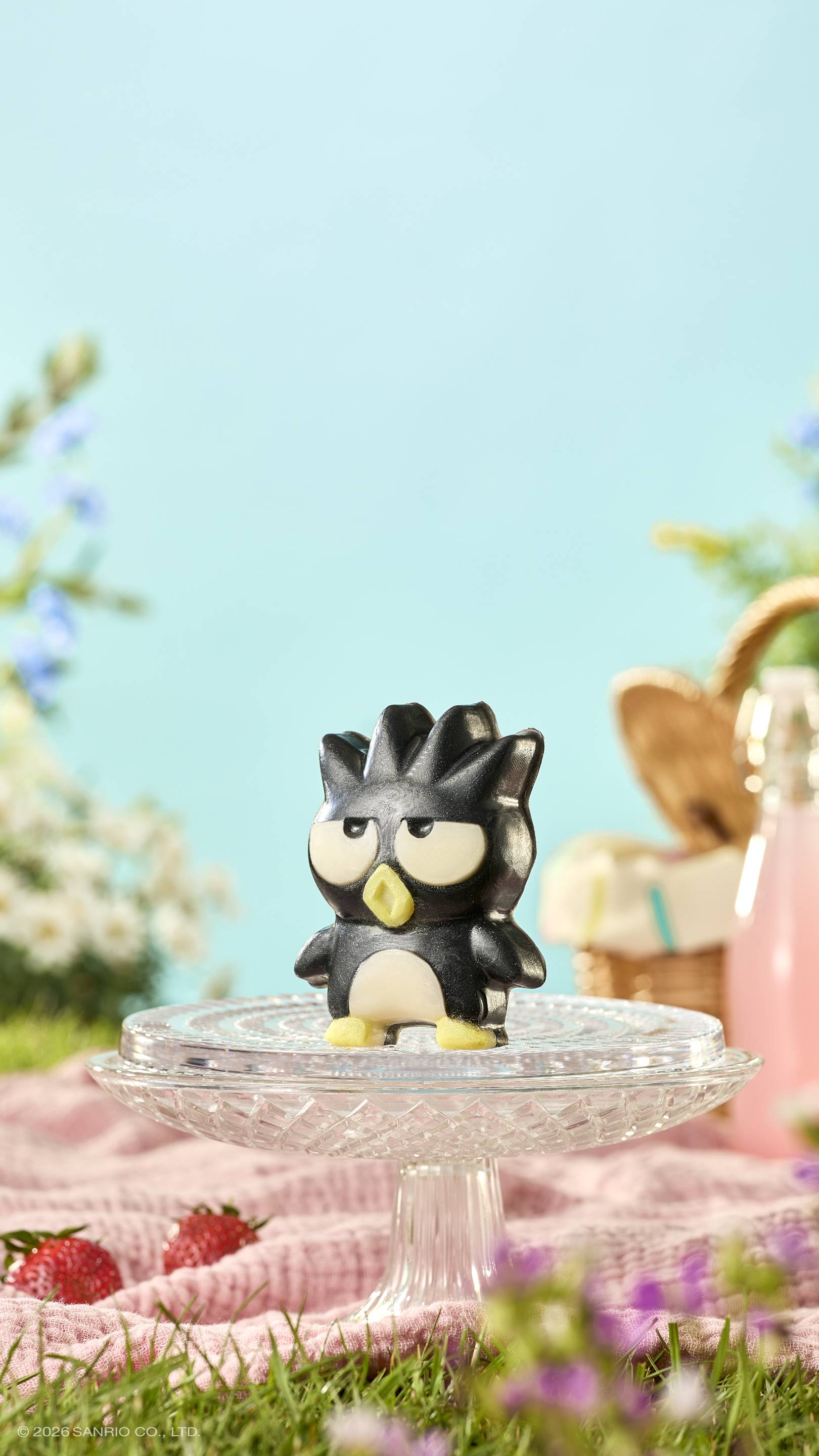 A black penguin-shaped soap with a grumpy expression sits on a glass cake stand. The sunny picnic setting features a pink blanket, strawberries, and a wicker basket against a soft blue sky. © 2026 SANRIO CO., LTD. appears at the bottom.