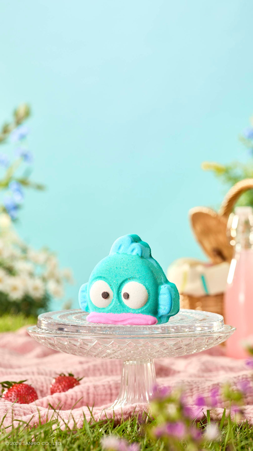 The teal, fish-shaped "Hangyodon" bath bomb sits on a glass cake stand in a sunny picnic setting. The background is a blue sky, with a wicker basket and a drink out of focus. A pink blanket and strawberries are scattered on the grass below.
