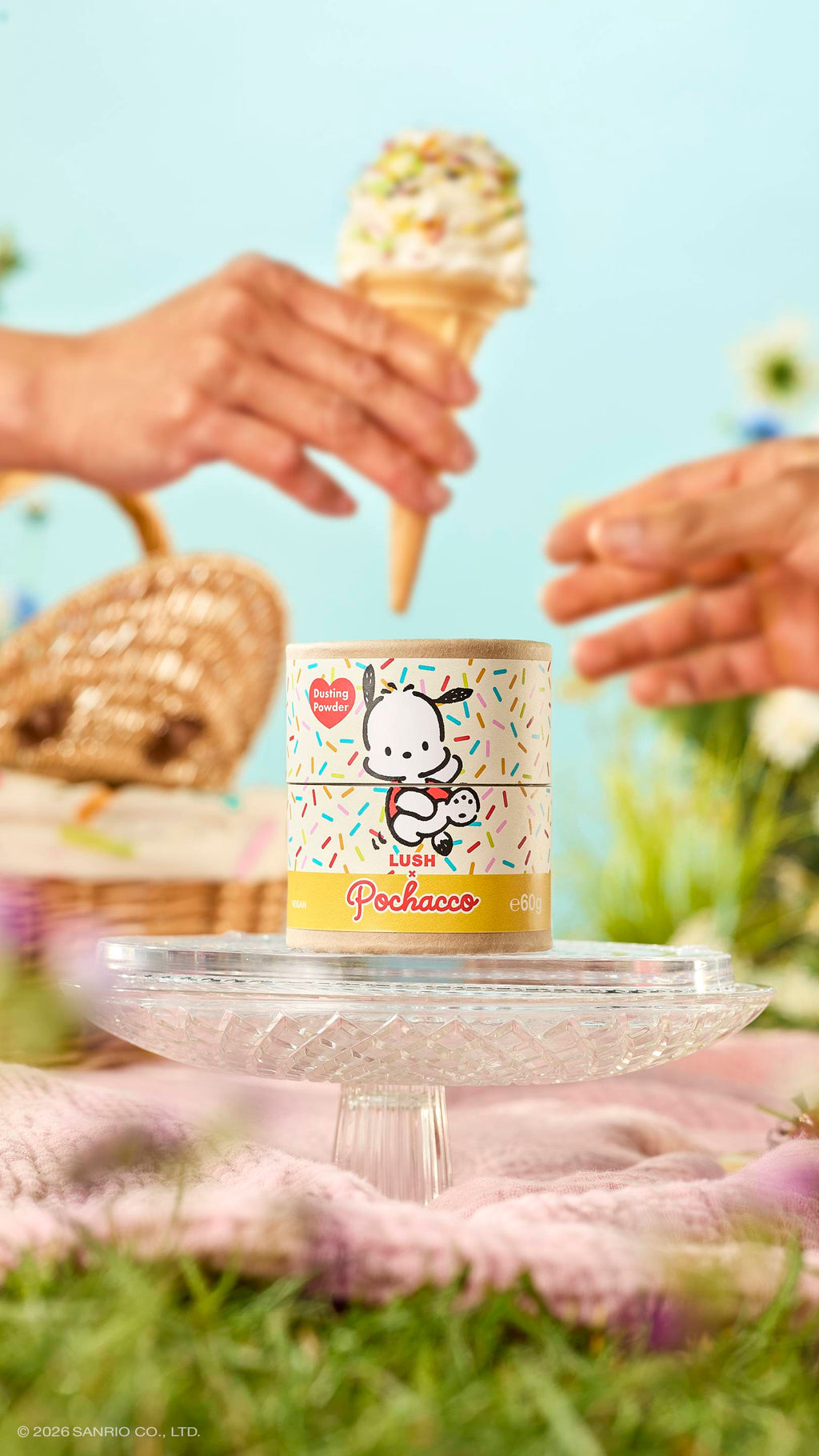 A cardboard tube of Lush x Pochacco dusting powder sits on a glass stand in a meadow. Above it, one hand passes a sprinkle-topped ice cream cone to another. The sunny picnic scene includes a wicker basket and a soft blue sky.