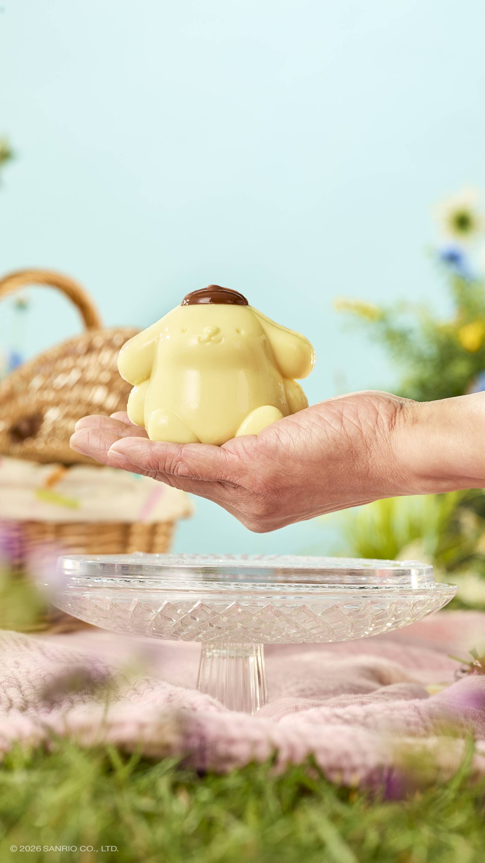 A hand holds a glossy, pale yellow "Pompompurin" shower jelly over a glass cake stand in a sunny meadow. The dog-like character wears a brown beret. A wicker basket and pink blanket sit on the grass under a blue sky.