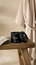 A slightly off-center shot of the black Super Milk Scented Candle tin, sitting open on a rustic, weathered wooden stool. The lid rests leaning on the tin, and a light-coloured towel and a beige bathrobe hang in the soft-focus background.