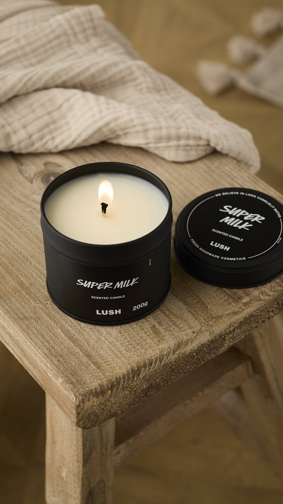 A close-up, slightly aerial view of the black Super Milk Scented Candle lit on a weathered wooden stool. The creamy white wax is melting around the bright wick, with the lid resting beside it and a beige cloth draped in the background.