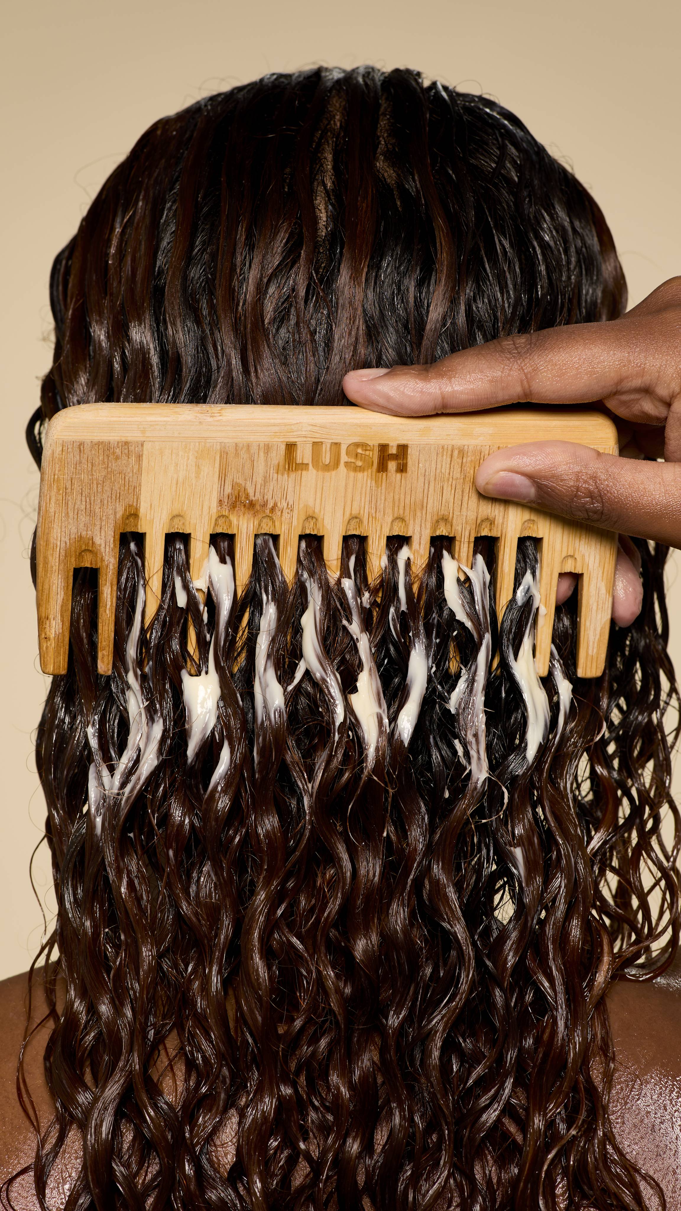 From behind, a hand uses a wooden Lush comb to detangle wet, dark, curly hair generously coated with a creamy Super Milk conditioner, creating beautiful, defined ringlets.