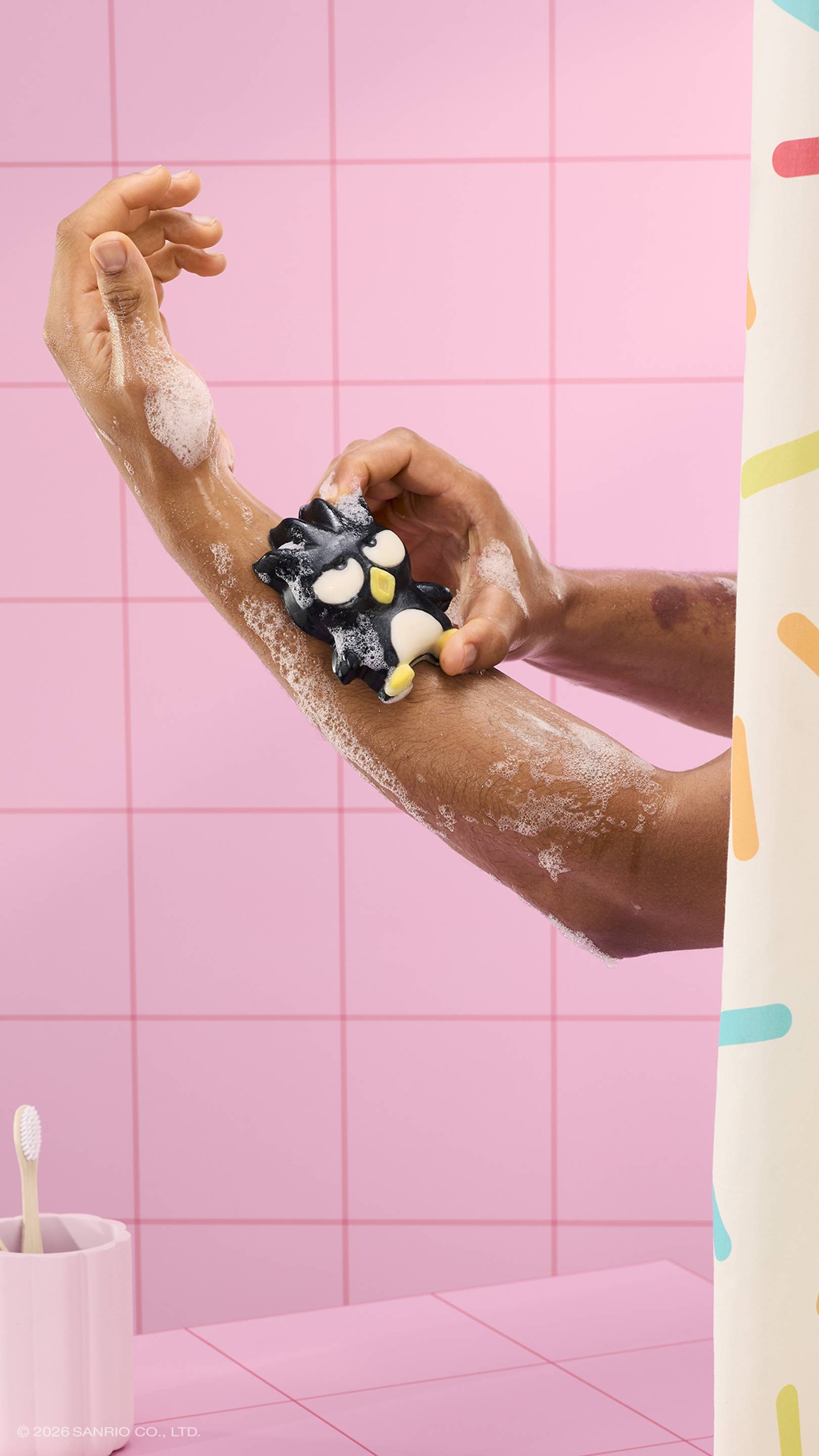 A person uses a black penguin-shaped "Bad Badtz-Maru" soap character to lather their arm in a pink tiled bathroom. The soap is covered in white bubbles, and a sprinkle-patterned shower curtain is visible on the right.
