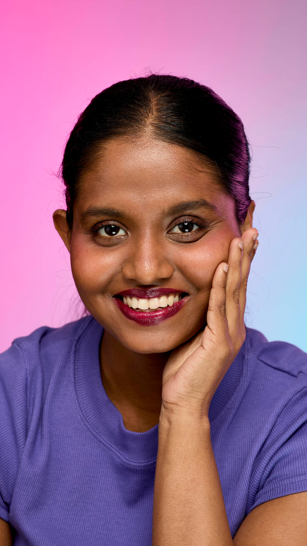 A smiling person with dark hair and skin, wearing a purple top, is shown from the chest up. They are wearing the Lush, deep plum Tartlet lip cream and have one hand touching the side of their face. The background is a pink and purple gradient.