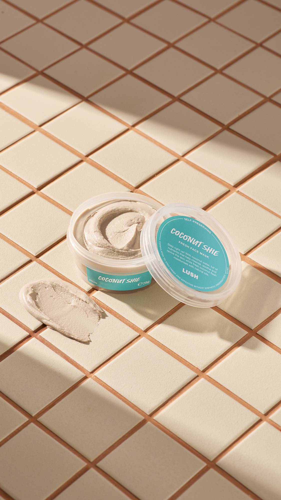 The image shows a clear, recycled plastic, open pot of Coconut Shie face mask with teal labels, the lid leaning on the side and a smeared swatch of the textured product on the cream-tiled floor beside the pot.
