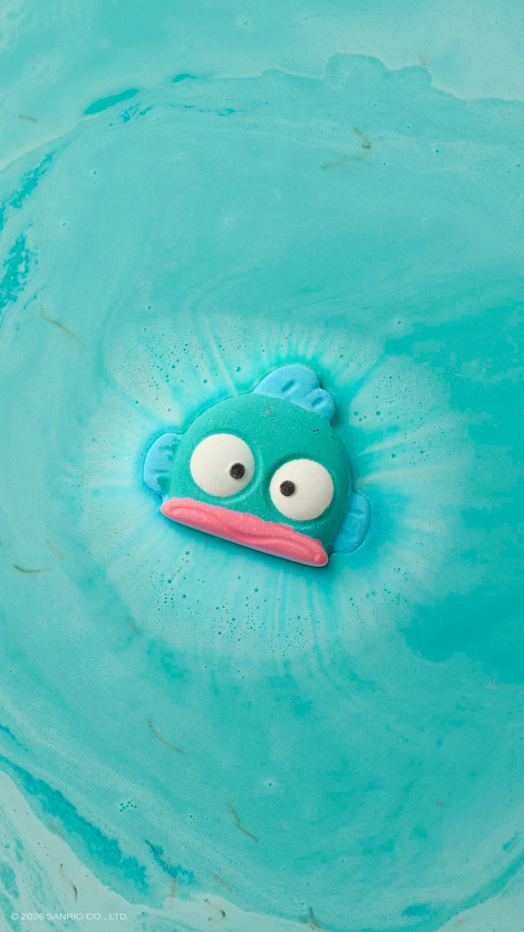 Hangyodon. A teal, fish-shaped bath bomb floats in a bath of turquoise water. It has large white eyes and a pink mouth, and is surrounded by a ring of white foam and bubbles as it dissolves.