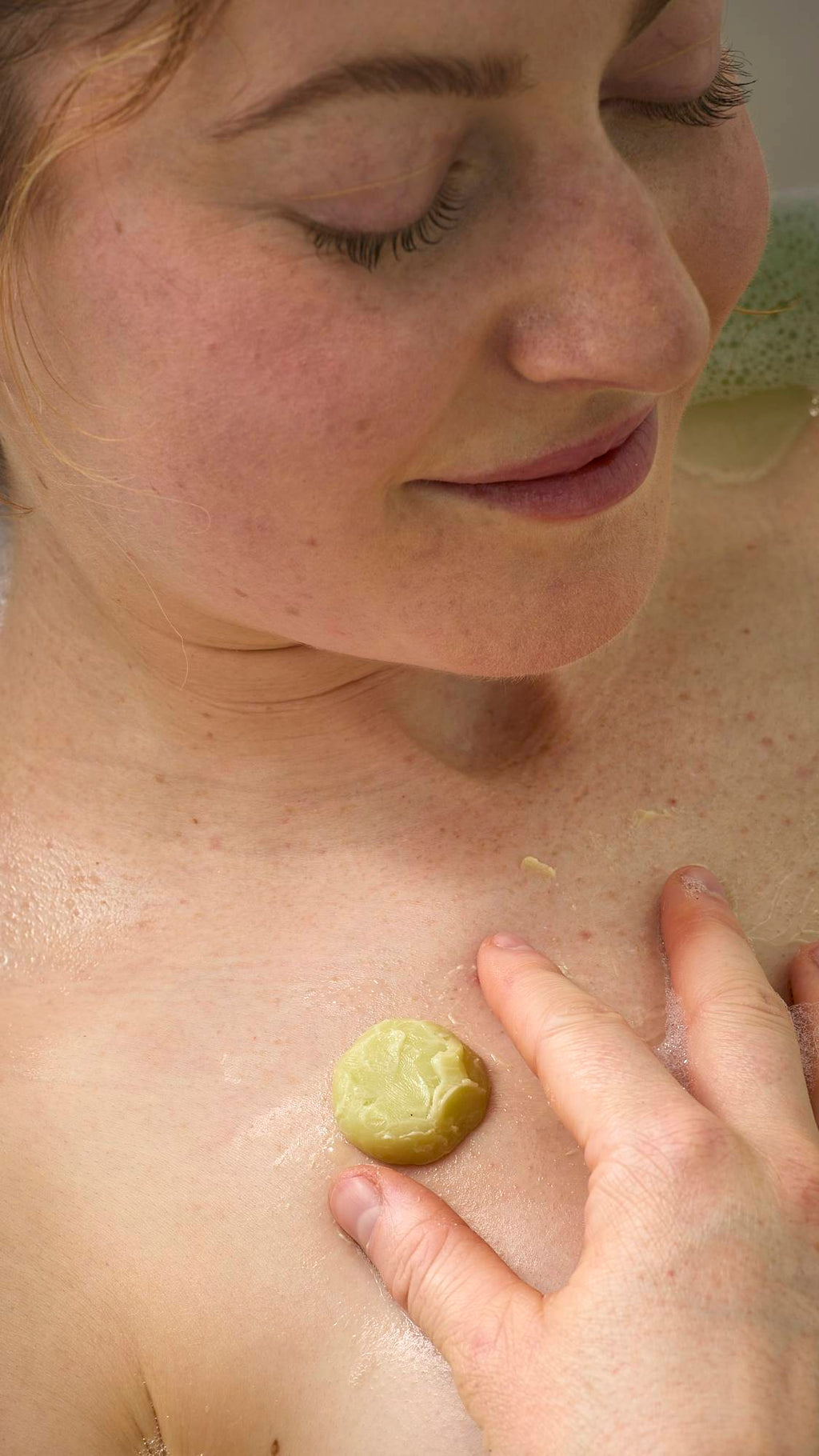 A smiling person relaxes in the bath, massaging the small, moisturizing melt from the Cold Bath Bomb onto their chest. The yellowish-green butter glides across their wet skin, offering a moment of soothing hydration.