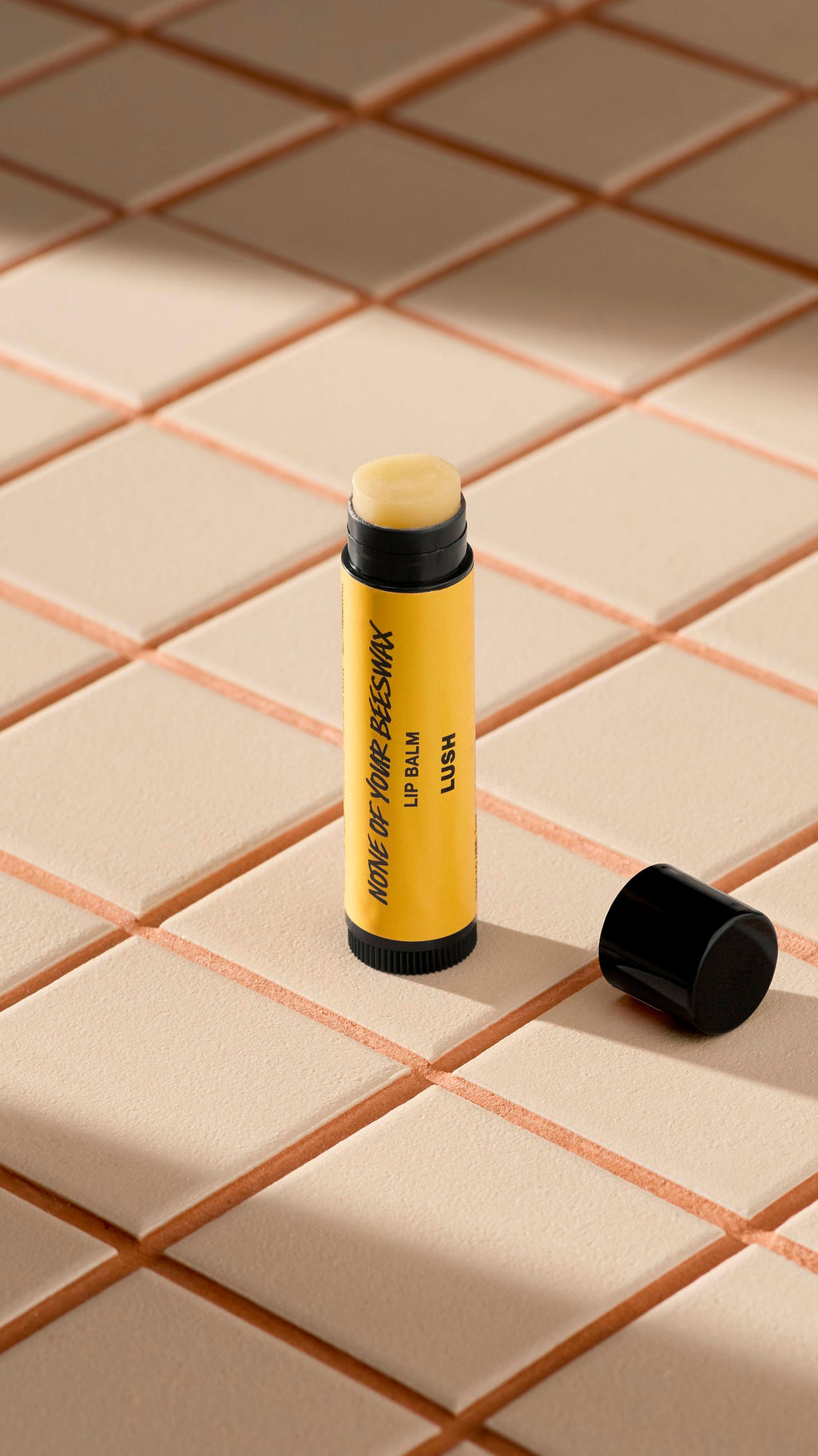 A yellow tube of Lush 'None of Your Beeswax' lip balm stands upright on light-coloured square tiles. The black cap is off, lying next to the base, revealing the pale, creamy balm inside. Soft shadows fall across the tiled surface.