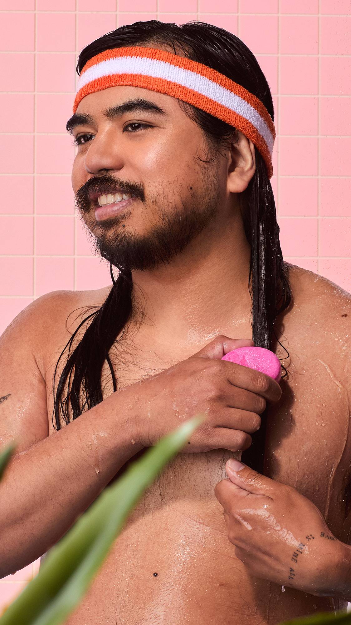 A smiling person wearing an orange-and-white headband glides a pink Lush "American Cream" solid conditioner bar down long, wet, dark hair in a shower with soft pink tiles.