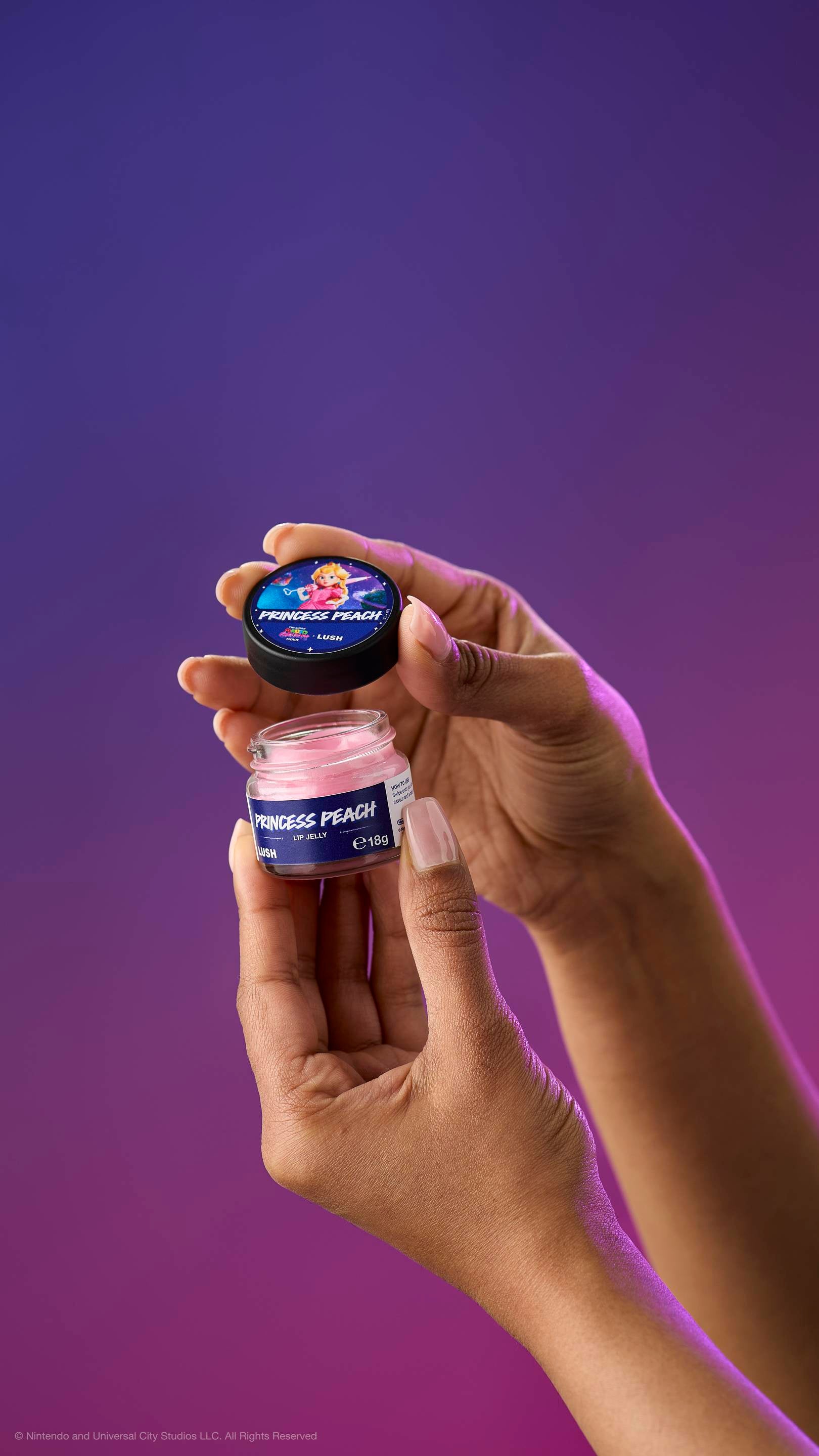 Two hands hold an open pot of creamy pink Princess Peach lip jelly and its lid against a purple-magenta gradient. The lid features Princess Peach holding a heart-shaped wand.