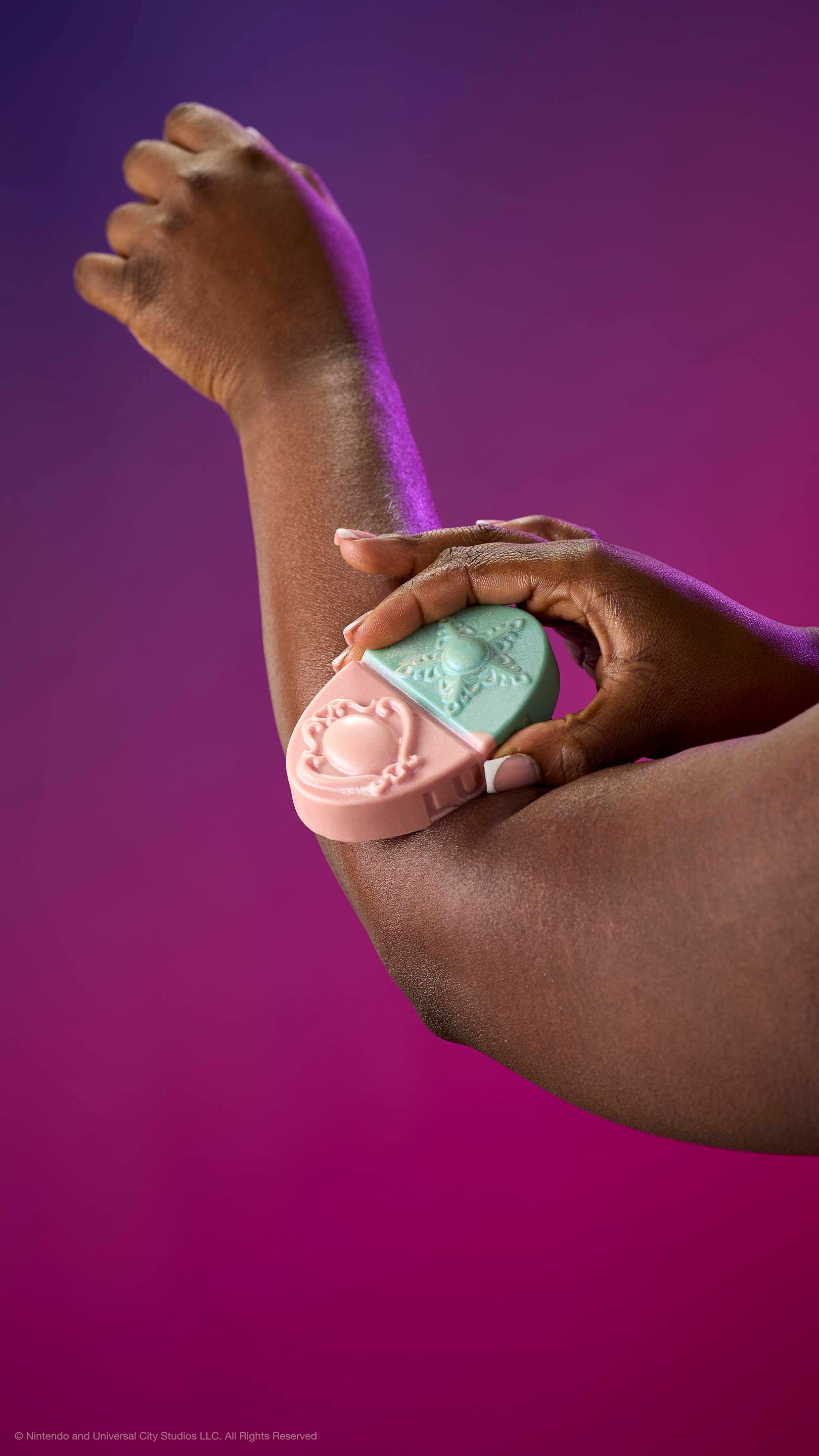 A hand glides the pink and teal Princess Power body balm over an elbow. Features embossed brooch and star designs. Against a purple-magenta gradient with plastic-free glitter.