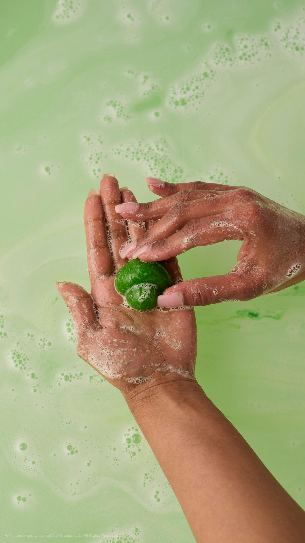 Hands hold a small, green mushroom-shaped soap over frothy, light-green bathwater.