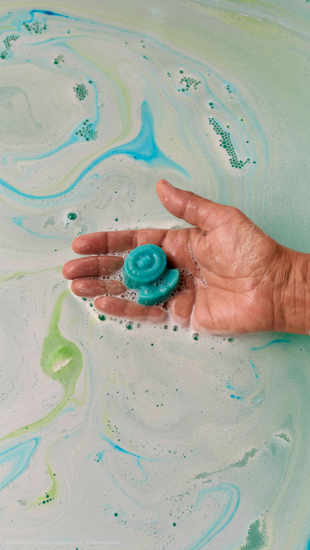 A hand holds a small, light-blue soap shaped like an Ice Flower over bathwater filled with swirls of white, light-blue, and green foam.