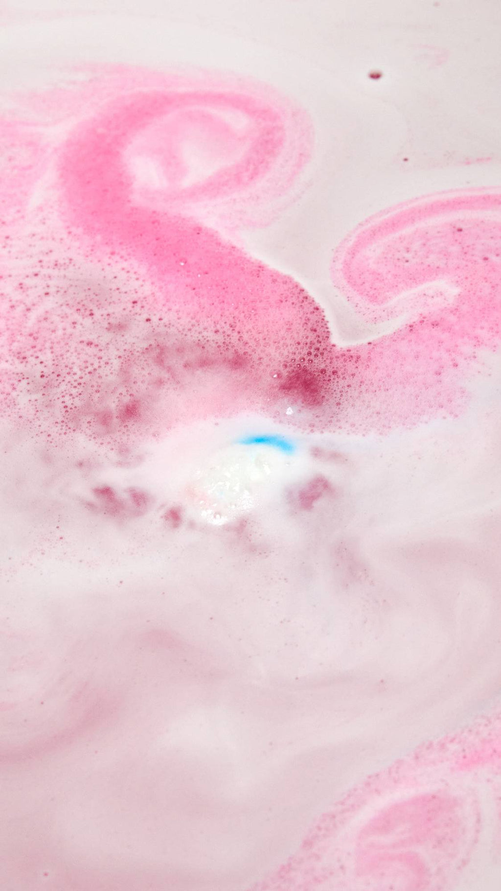The American Cream bath bomb is slowly dissolving in the bath water giving off a pretty pink and white rippled foam across the surface with a small speck of blue in the centre.