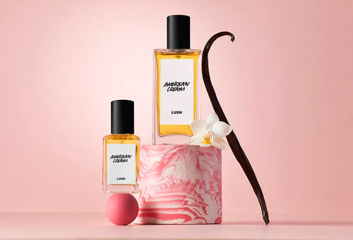 Two perfume bottles balance on a pink ball an cylindrical podium. Next to it is a pink and white flower and a vanilla pod.