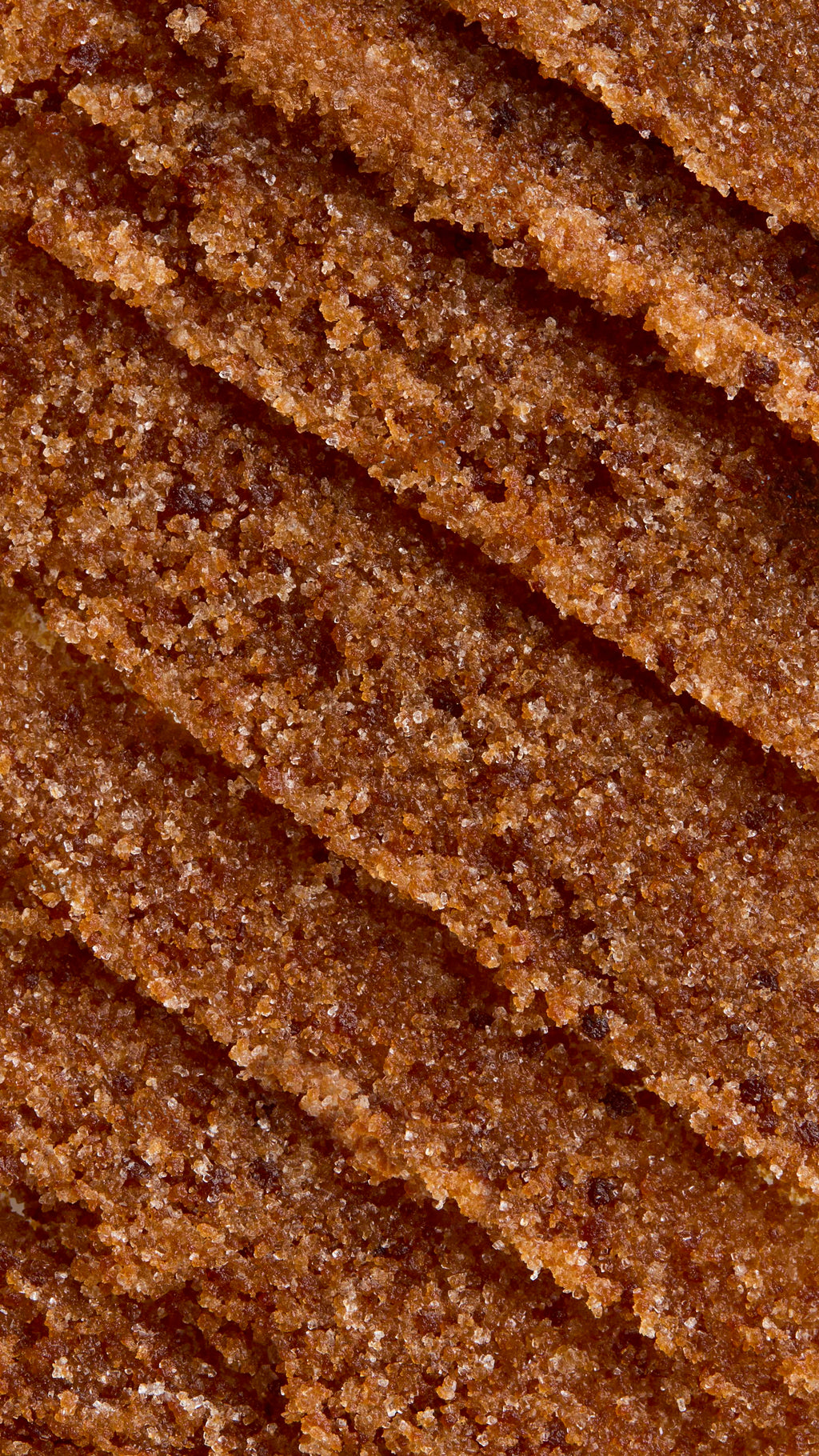 A close-up, aerial view of textured, thick caramel-coloured Buzz lip scrub. The texture is rich and gritty, arranged in several diagonal, overlapping swoops that create ridges and valleys.