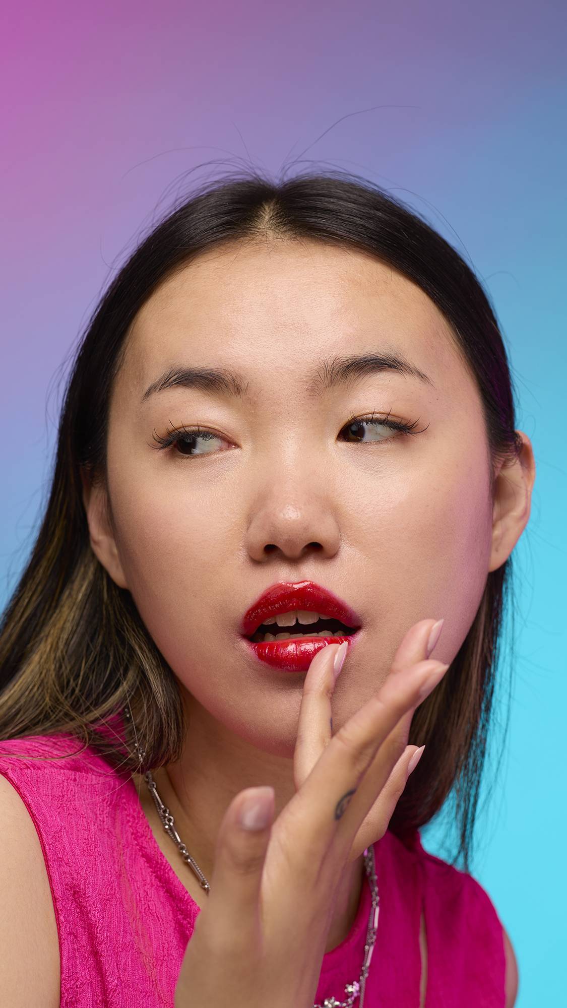 The model has long, dark hair and is on a pastel pink, blue and purple hue background. Their hand is by their face adjusting the juicy coating of blushed-red tint on their lips created by the Cherry lip jelly.