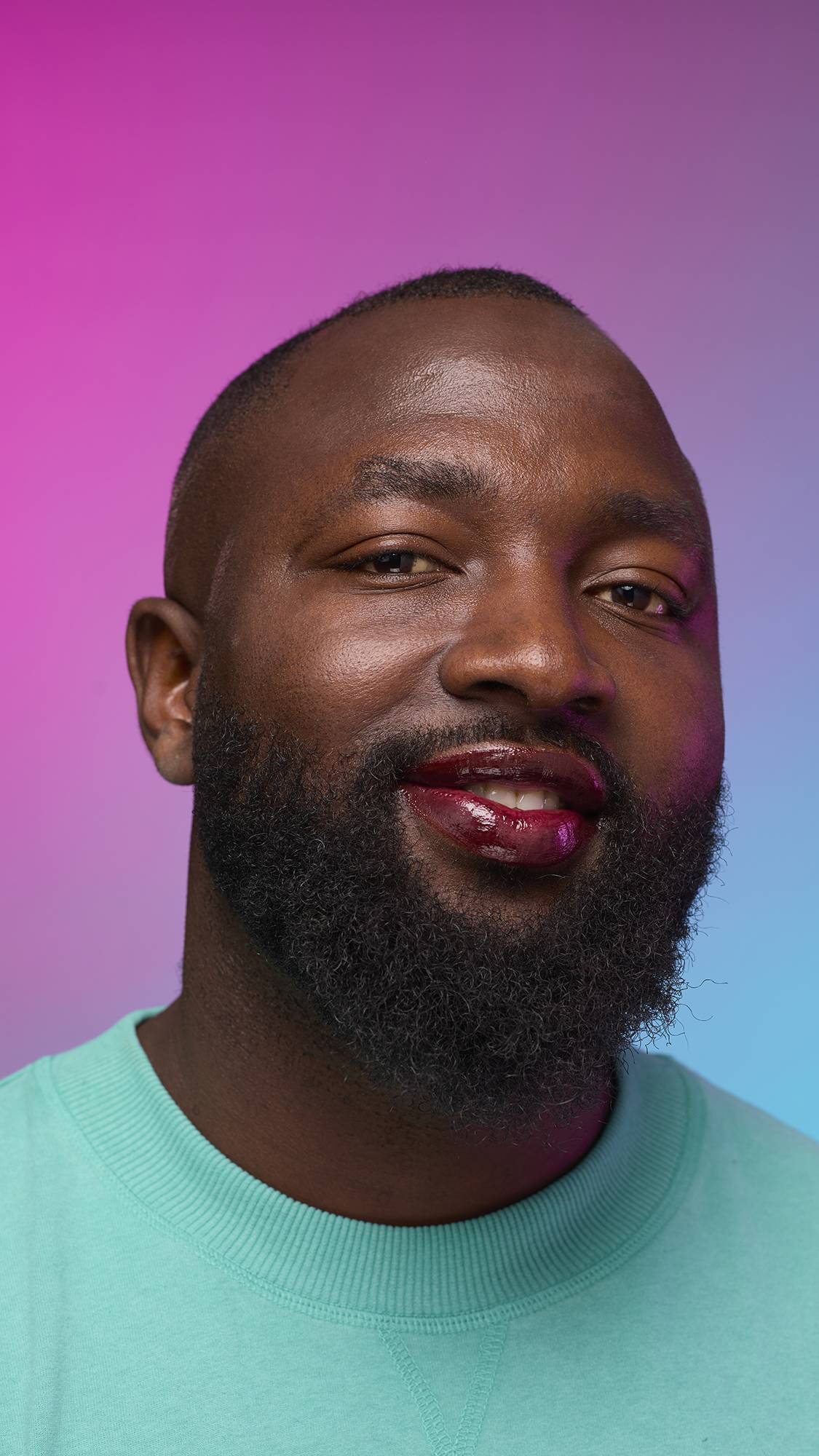The model is on a pastel pink, blue and purple hue background in a vivid teal jumper. They are glancing away with the focal point being the juicy coating of grounding, earth-toned tint on their lips created by the Cola lip jelly.