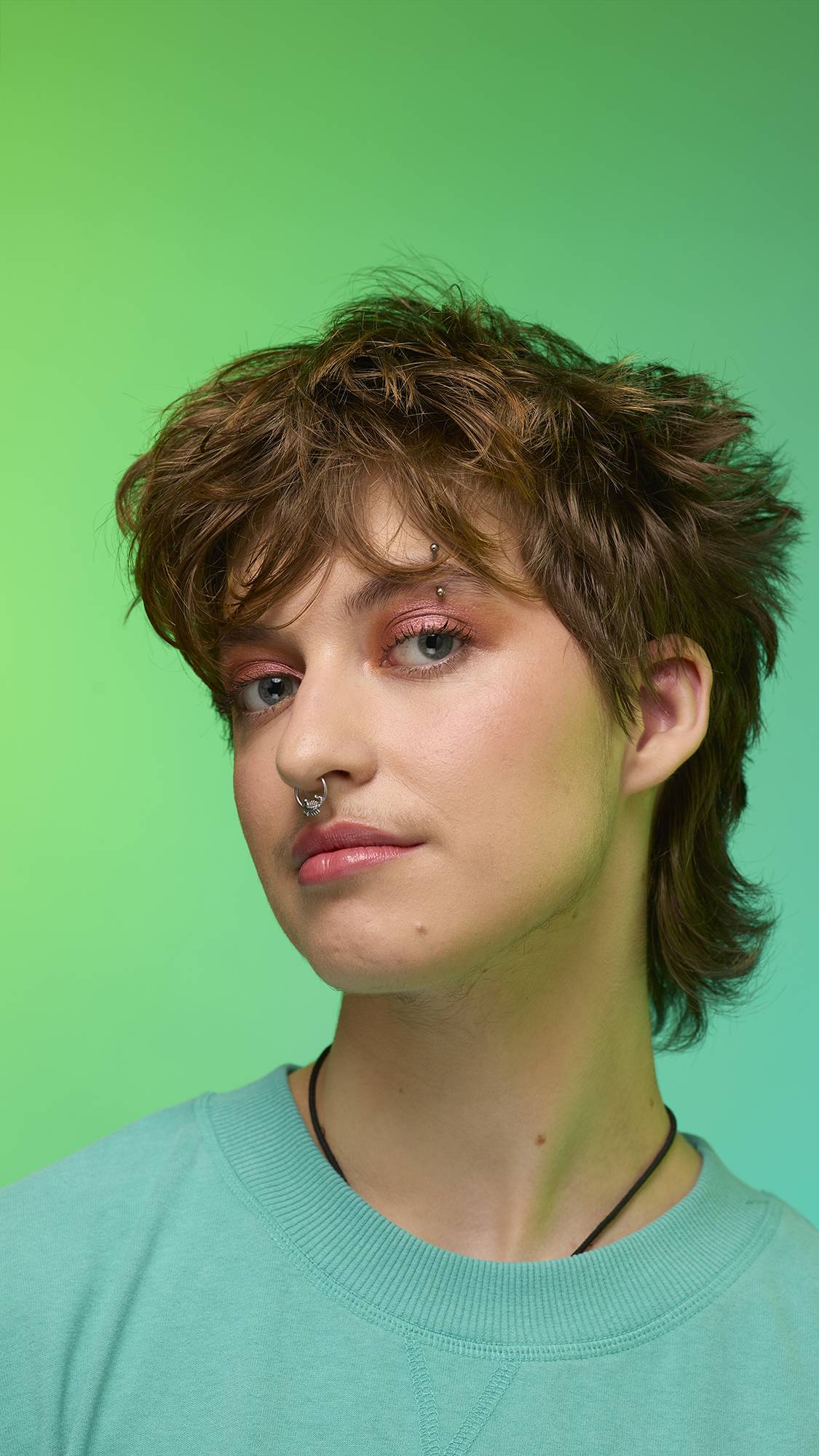 The image shows a close-up of the model's face on a pale blue-to-green gradient background. The model's make-up is done with the focal point being a shimmering, metallic-bronze eyeshadow made by the Comet Glitter Crumble.