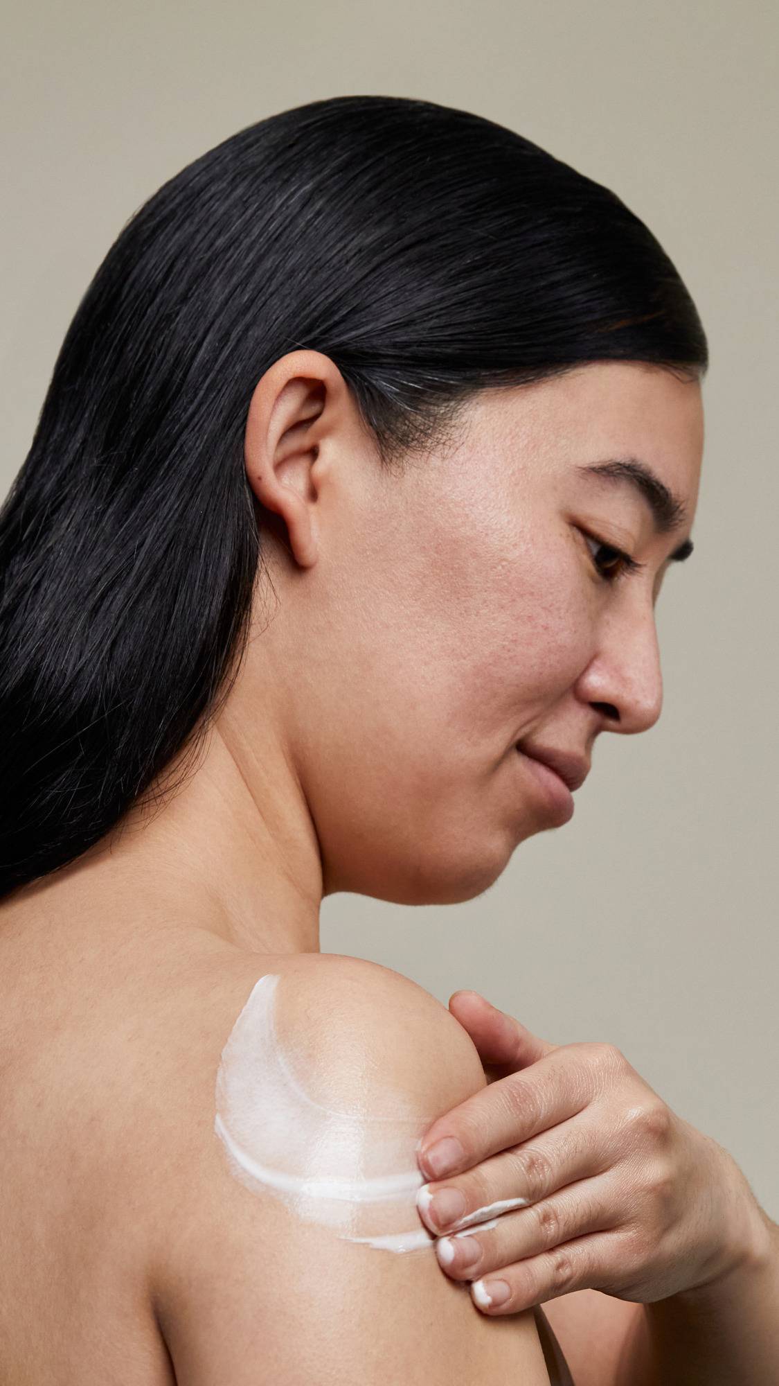 The image shows a side view of the model. Their dark hair lays straight down as they apply the body lotion in circular movements over their shoulder.