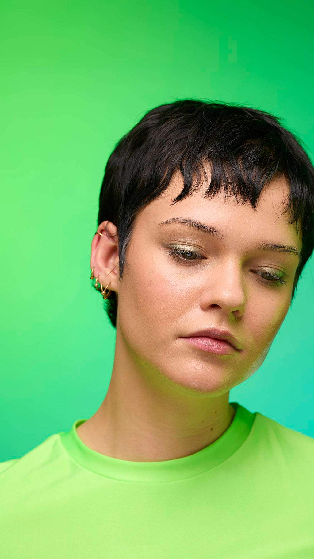 The image shows a close-up of the model's face on a pale blue-to-green gradient background. The model's make-up is done with the focal point being a subtle, gunmetal-brown, shimmering eyeshadow made by the Eclipse Glitter Crumble.