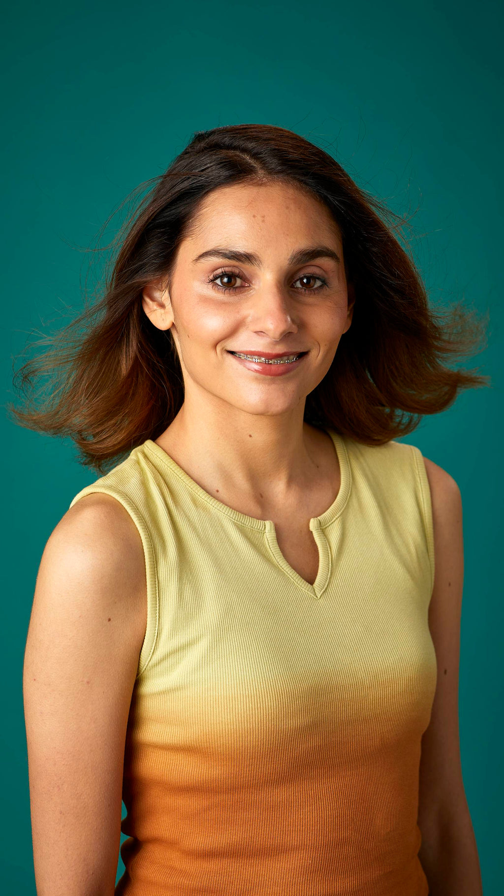 The model is on a teal background with a contrasting, yellow and orange gradient tank top. They have thick, shoulder-length hair which looks clean and healthy and is being fanned with air.