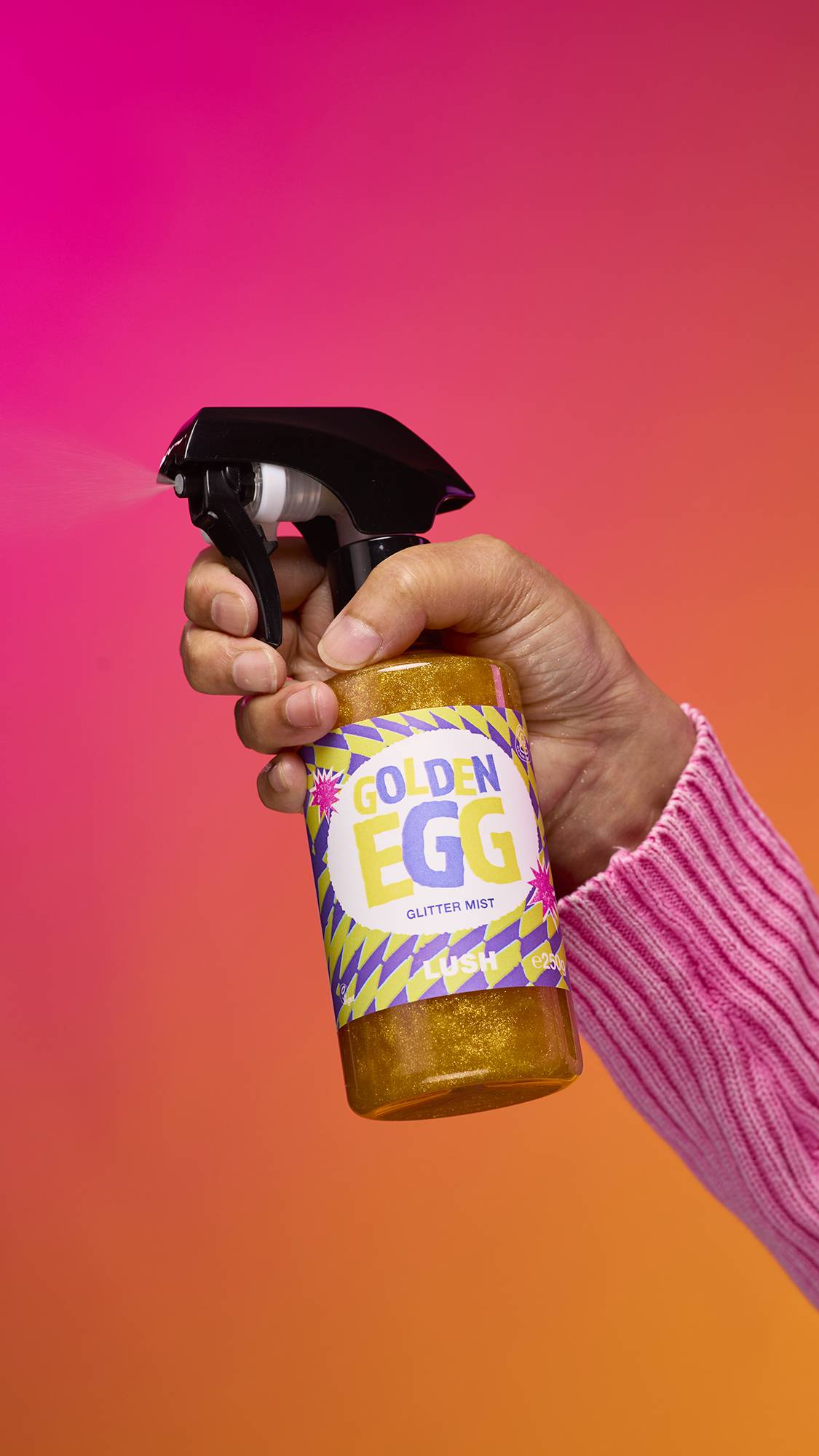 An arm is reaching from the right-hand side. The hand holds a transparent, cylindrical plastic spray bottle with golden, shimmering liquid and a black top. A colourful label reads "Golden Egg Glitter Mist" on a pink-to-orange gradient background.