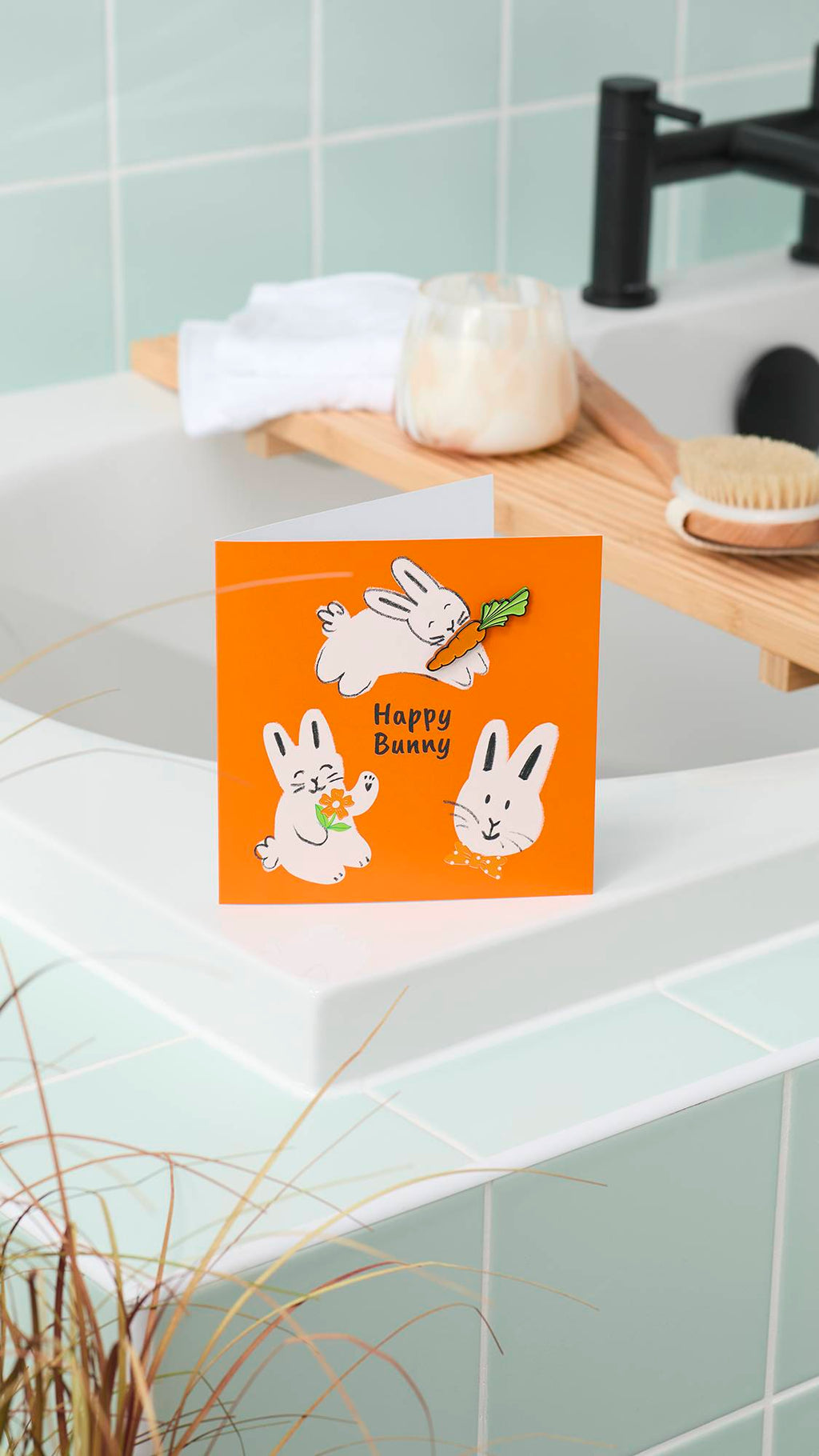 An orange "Happy Bunny" greeting card stands on the edge of a white bathtub. In the background, a wooden bath tray holds a candle and brush, set against mint-green wall tiles and a black tap.