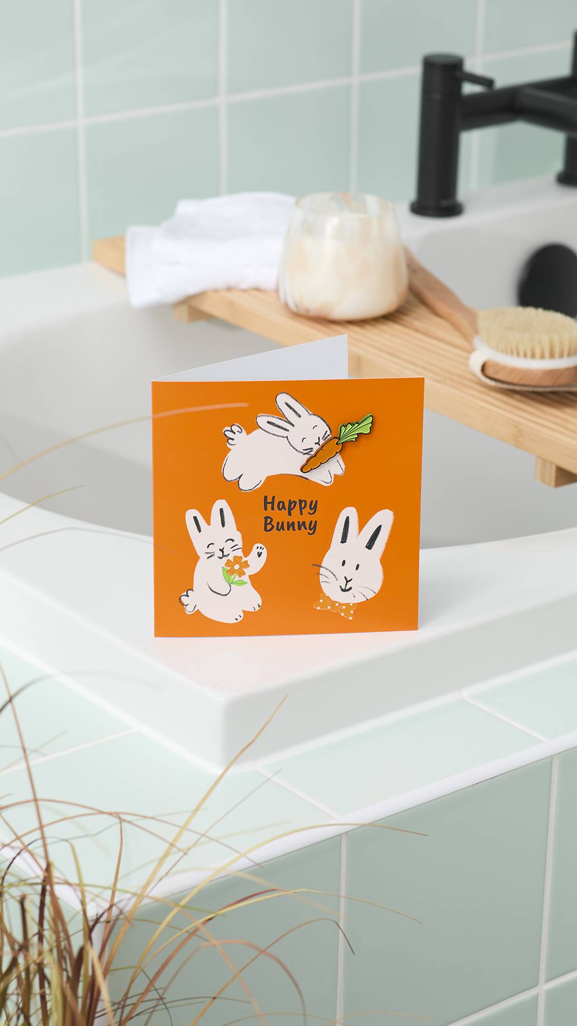 An orange "Happy Bunny" greeting card stands on the edge of a white bathtub. In the background, a wooden bath tray holds a candle and brush, set against mint-green wall tiles and a black tap.