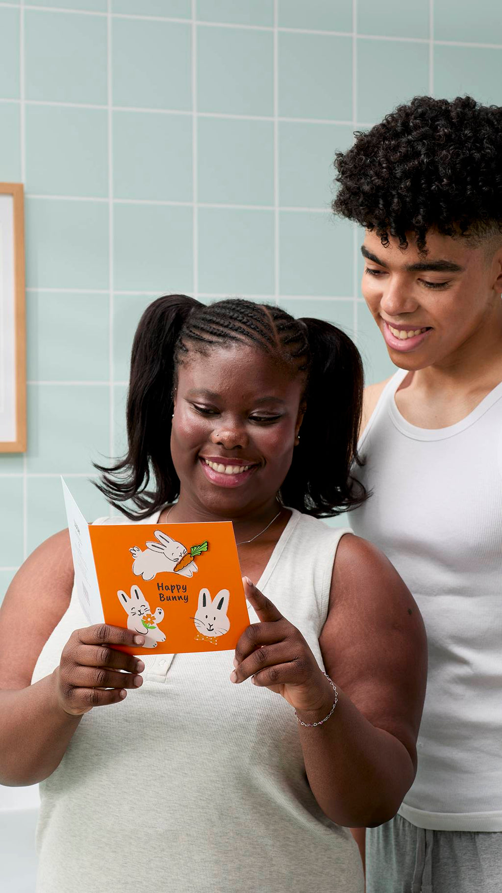 Two people smile while looking at an orange "Happy Bunny" greeting card held by a model with their hair in bunches. They are indoors against a backdrop of pale mint-green wall tiles.