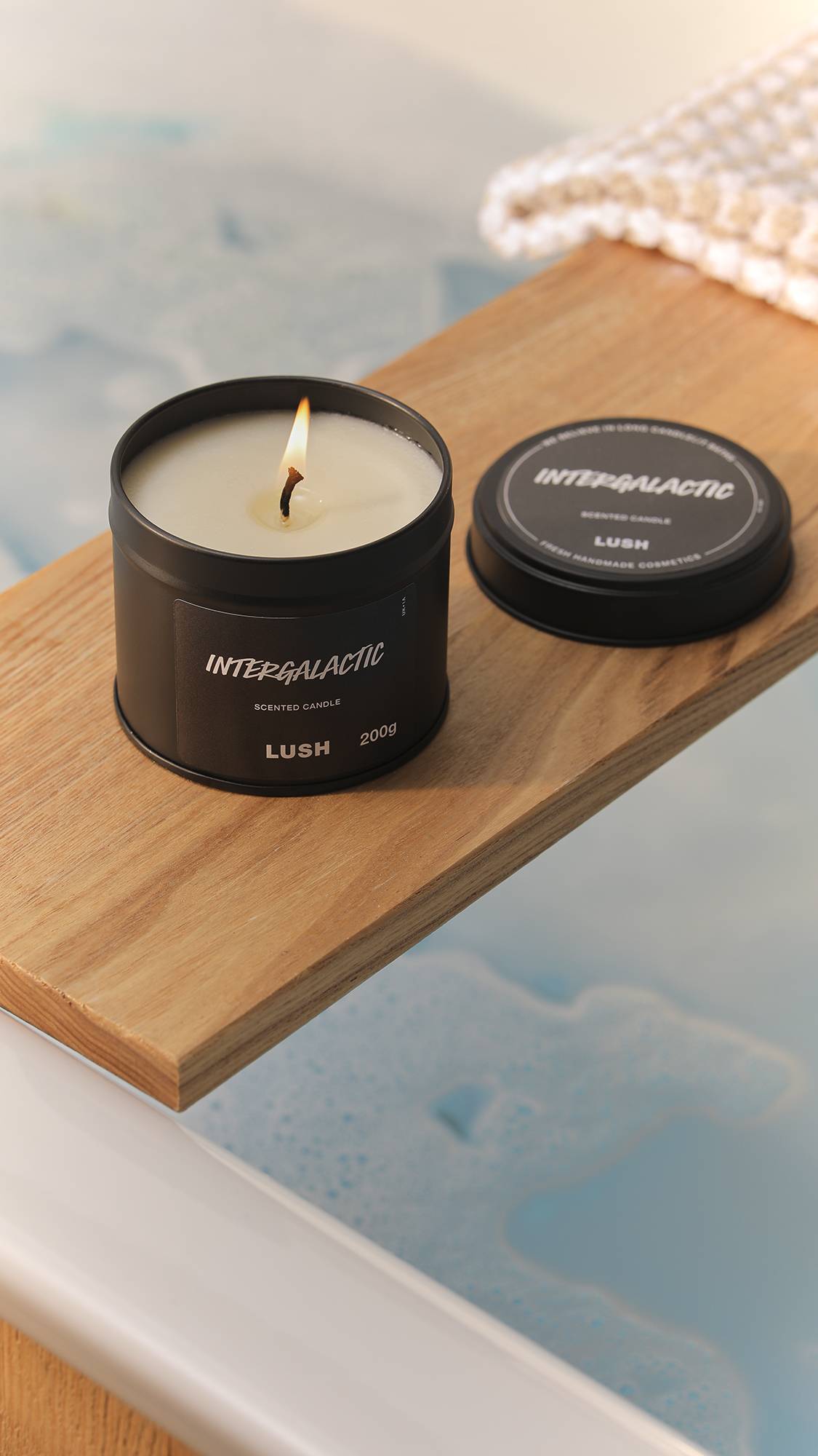 The image shows a bright, blurred bathroom scene. The black tin Intergalactic candle has a lit wick and is sitting on a wooden bath caddy laid over a filled bath.