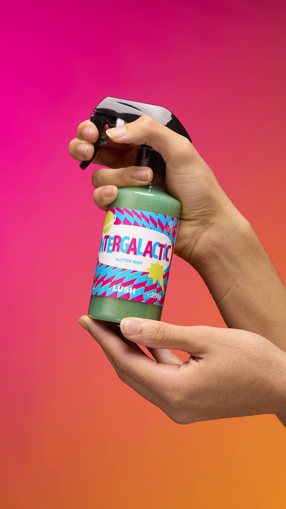 An arm is reaching from the right-hand side. The hand holds a transparent, cylindrical plastic spray bottle with teal, shimmering liquid and a black top. A colourful label reads "Intergalactic Glitter Mist" on a pink-to-orange gradient background.